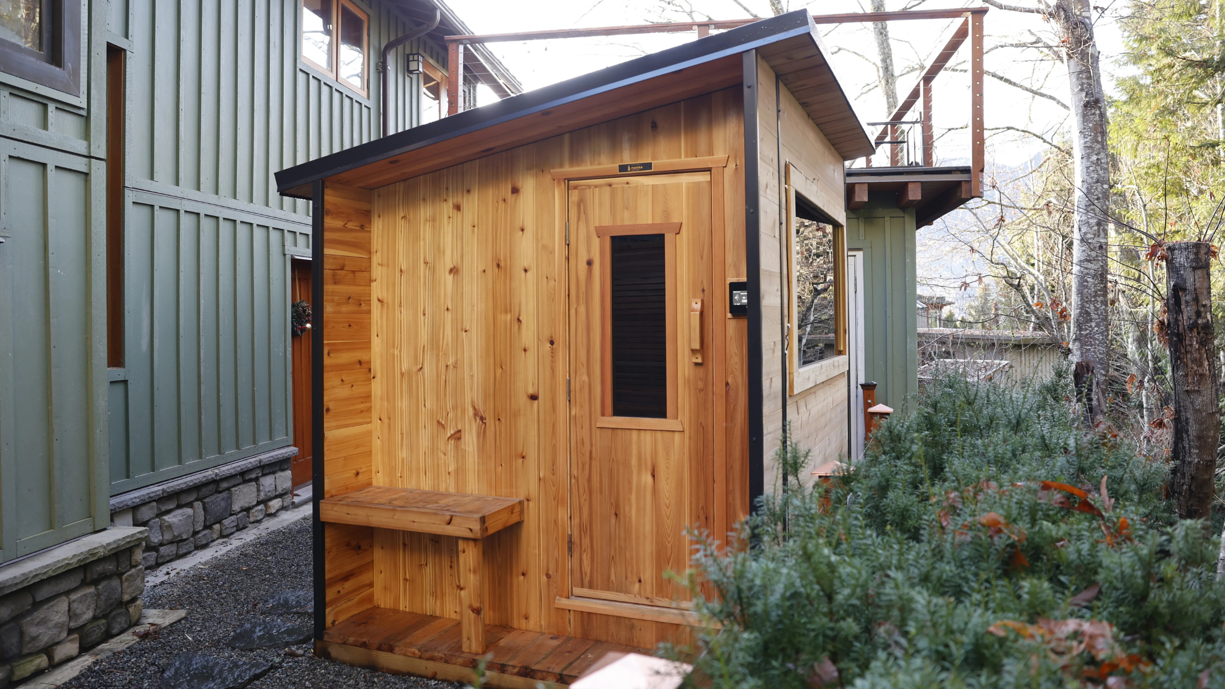wooden sauna with slanted roof and large window outside next to building and plants, best home sauna with a view