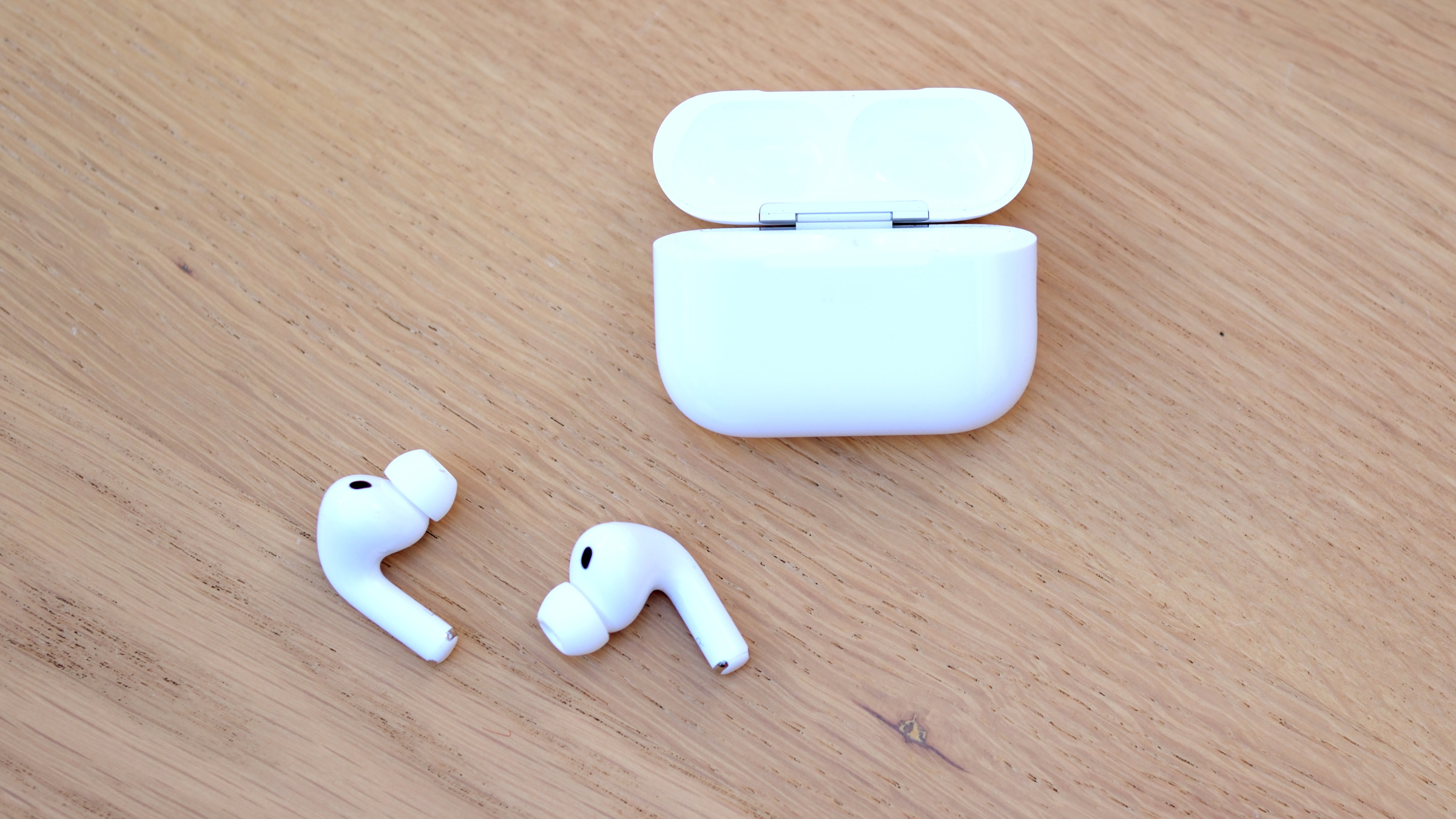 white Apple AirPods Pro 3 earbuds next to open charging case on wooden surface