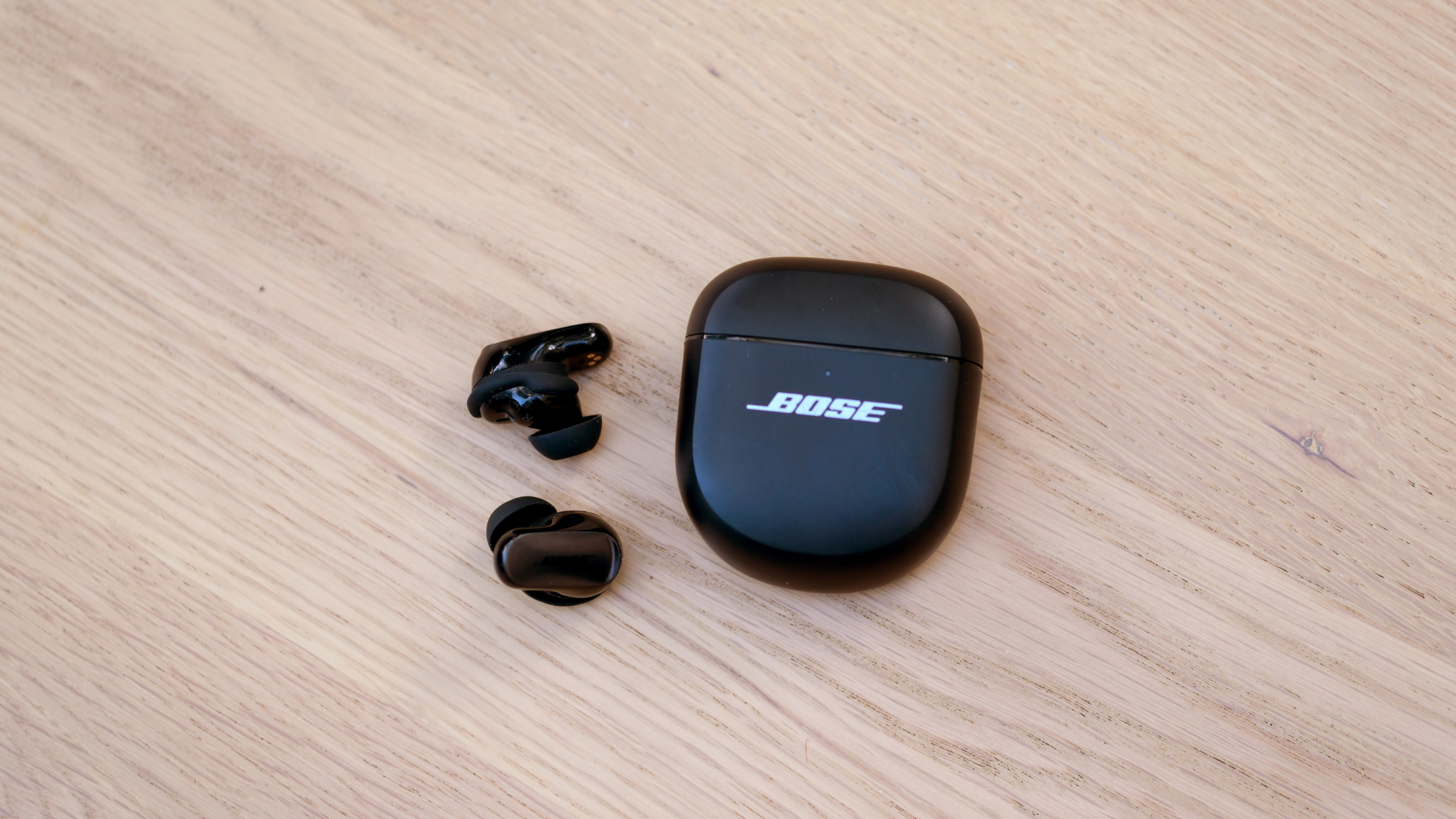 Bose QuietComfort Ultra (2nd Gen) black earbuds next to case on wooden surface, earbuds with best bass