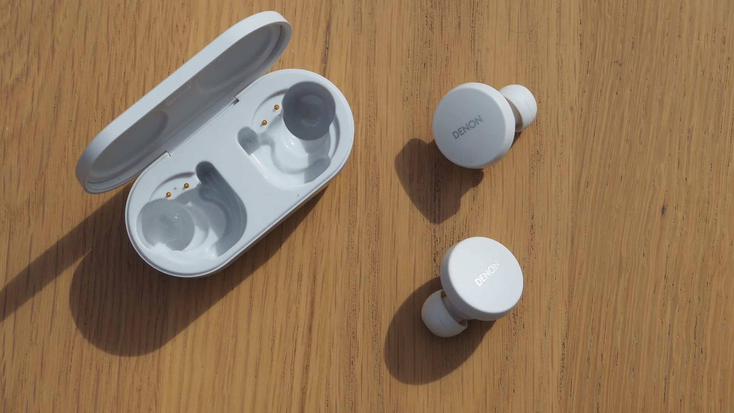 white Denon PerL True Wireless Earbuds next to charging case on wooden surface