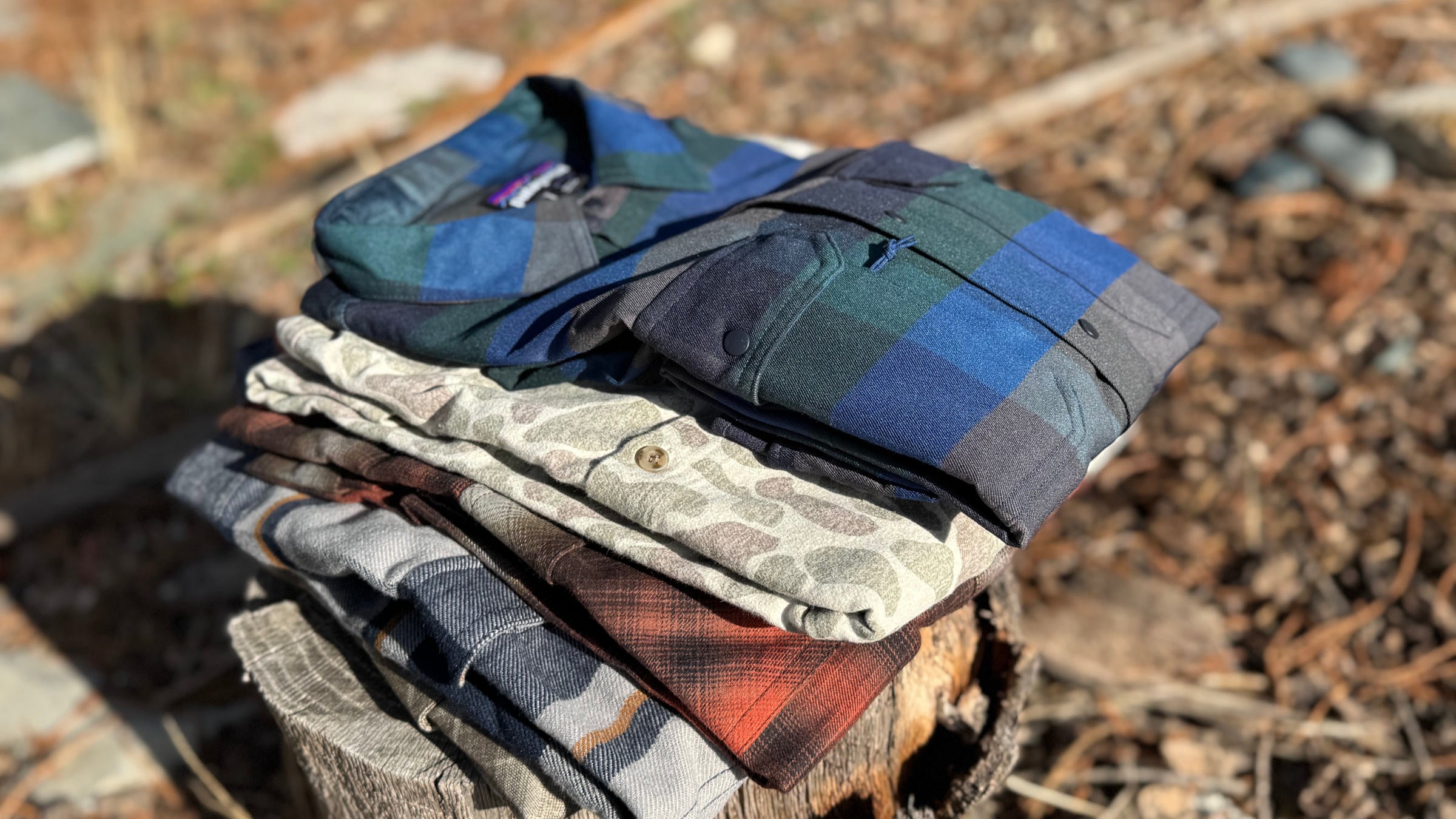 A stack of editor-tested picks for the best flannel shirts for men, including Patagonia and camouflage styles, displayed on a wooden stump in a forest setting.