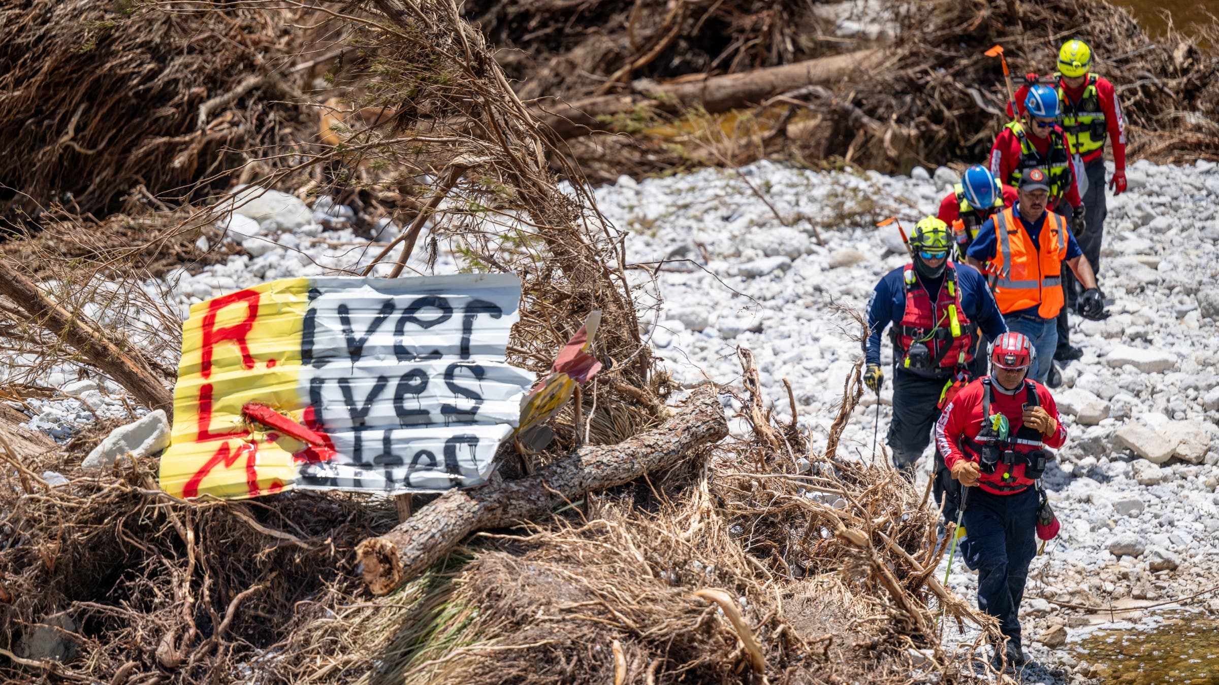 Search and Rescue workers crossing a flooded river in front of a sign that says, "River Lives Matter."