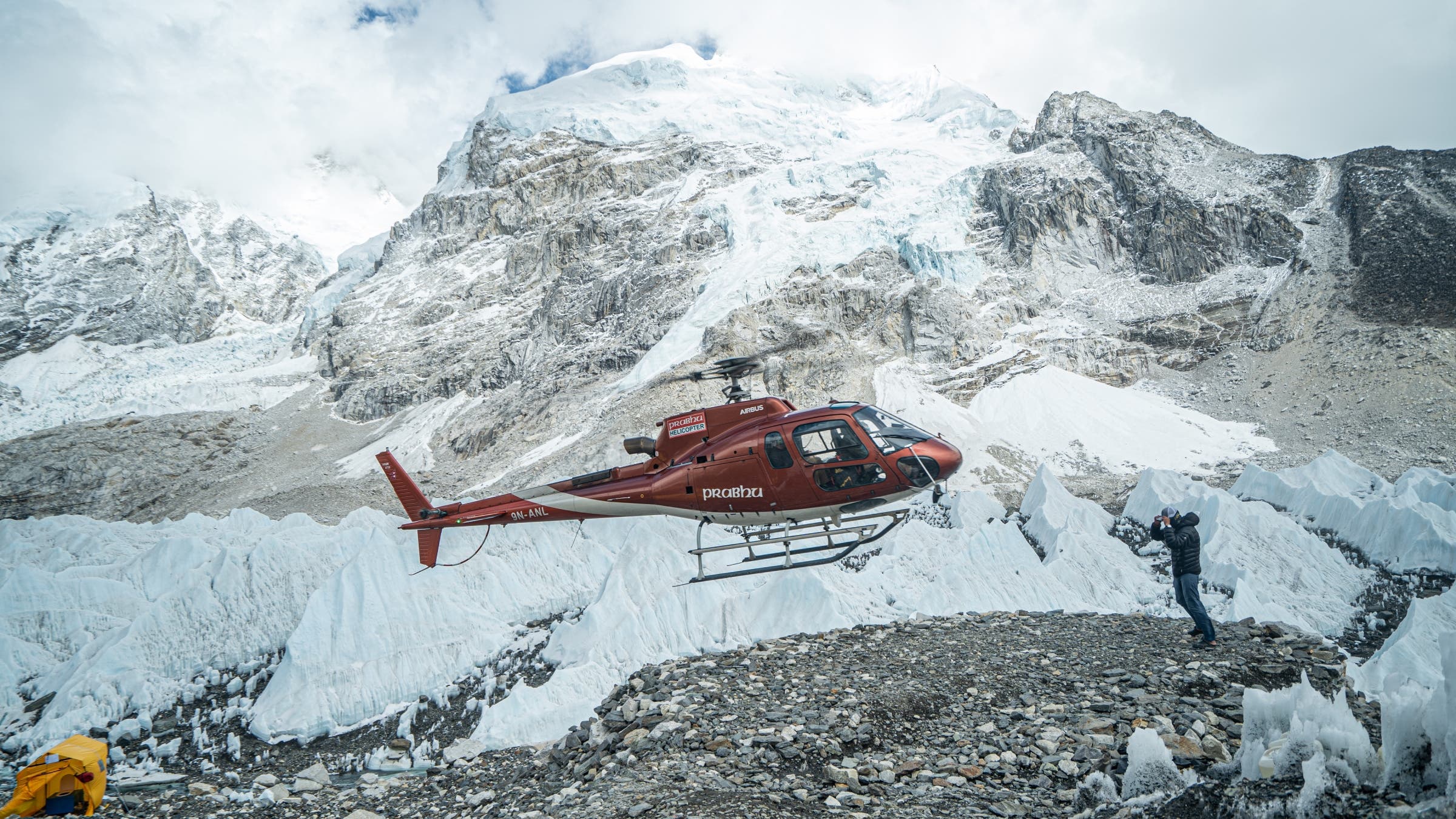 A helicopter hovers over a rock clearing with Mount Everest behind.