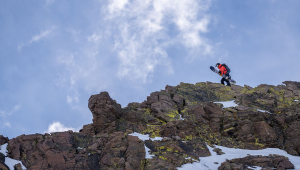 Snowboarder walks along rocky ridgeline with splitboard.