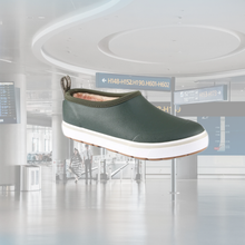 A Grundéns Deck Boss Shorty Sherpa boot in forest green featuring a white midsole and warm fleece lining, superimposed over a blurred background of a modern airport terminal.