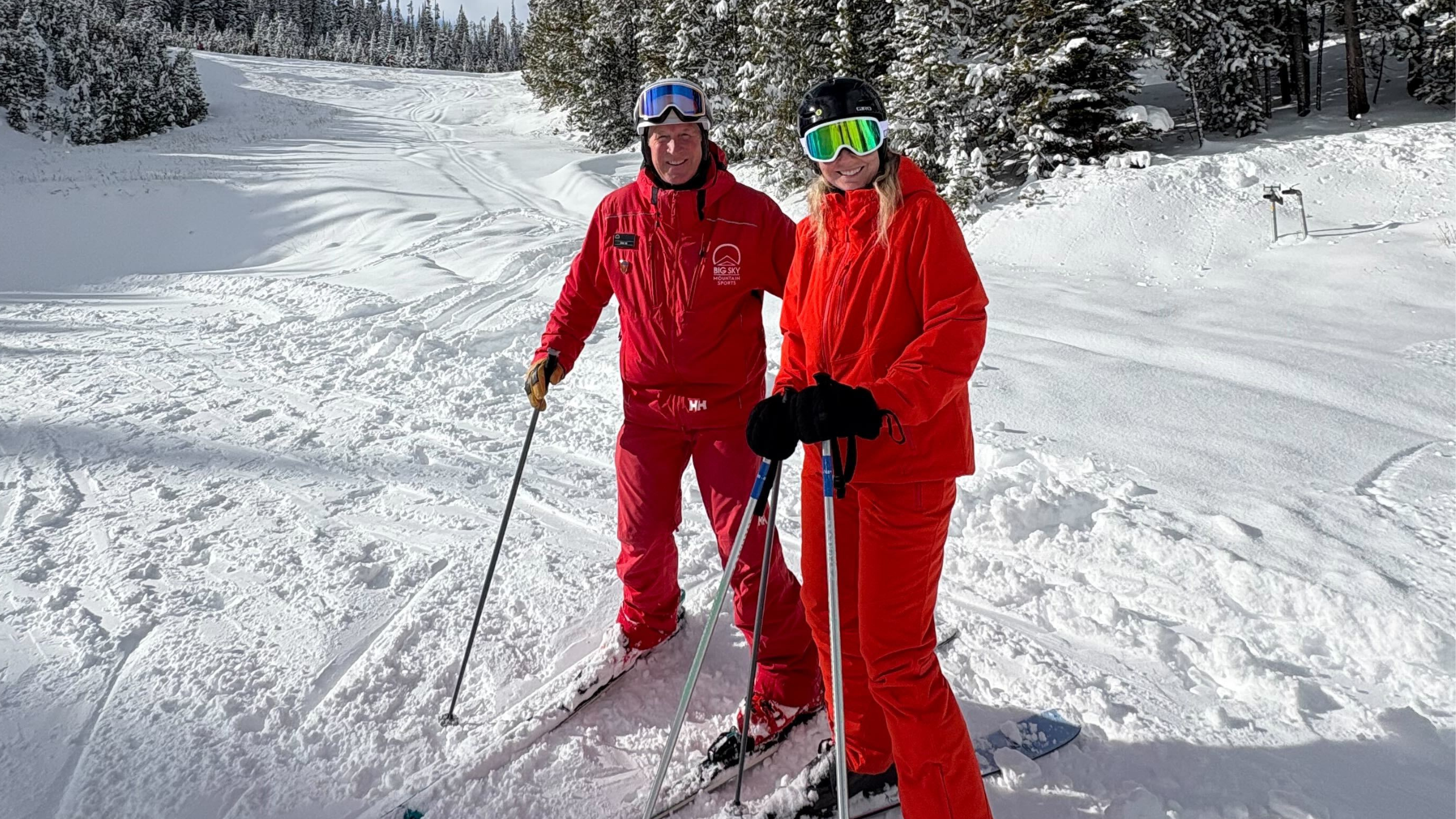 one man and one woman smiling at the camera in ski gear on a snowy mountain