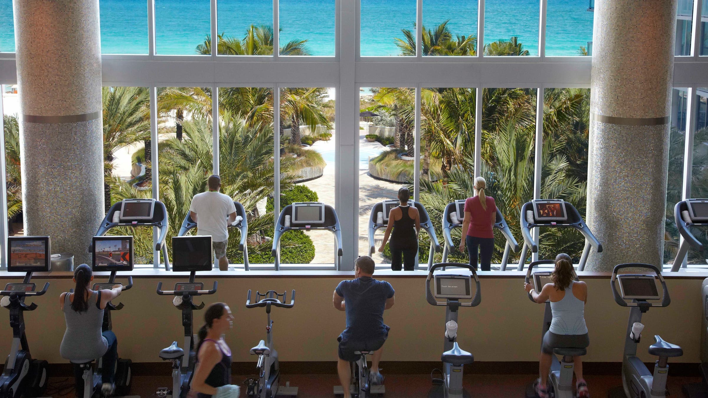 Hotel guests workout on cardio equipment with a beachfront view.