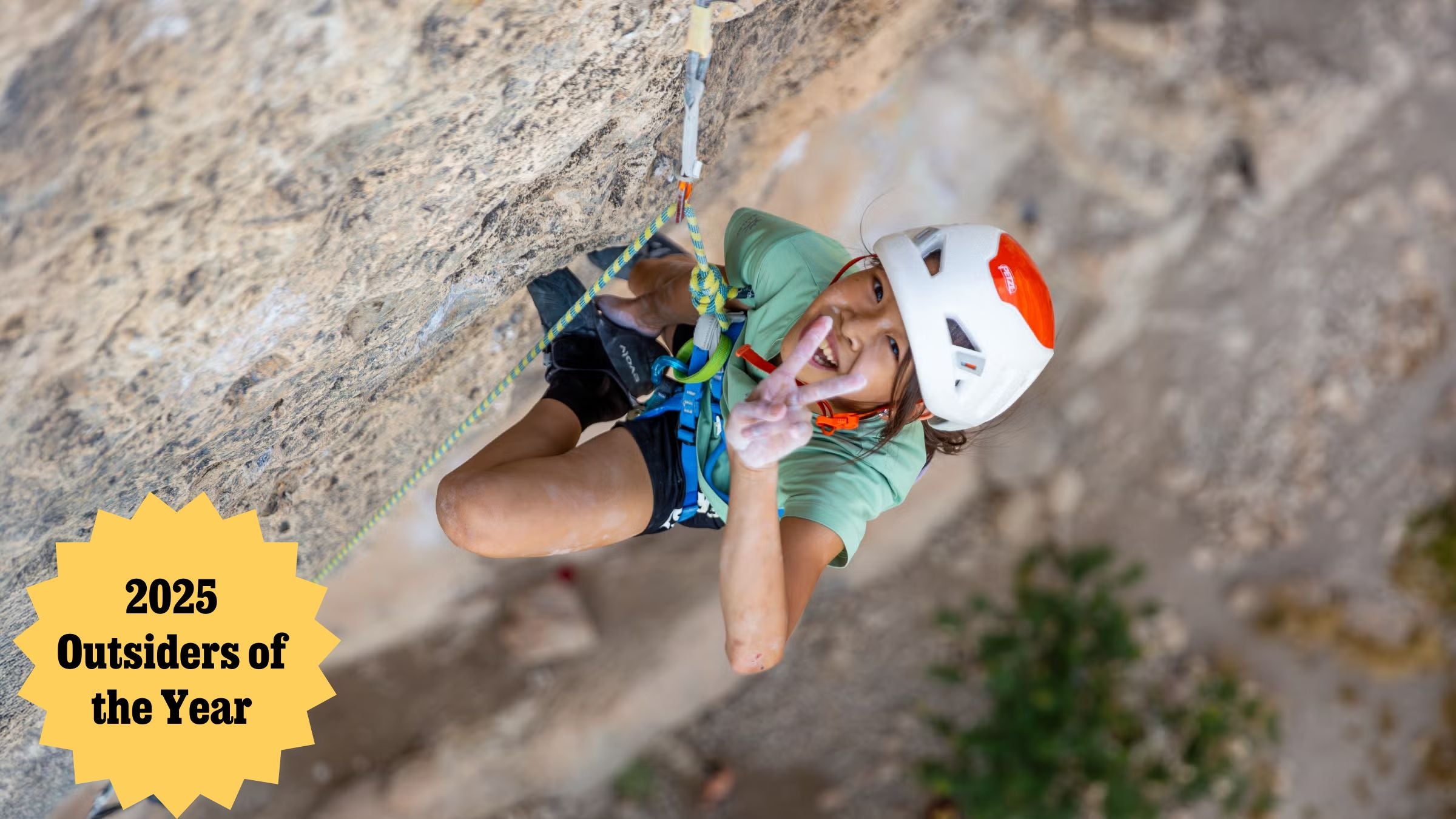 A girl gives a peace sign while hanging from a cliff.