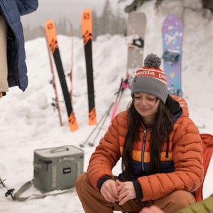 girl wearing cotopaxi jacket and patagonia beanie in the snow with skis and a cooler behind her