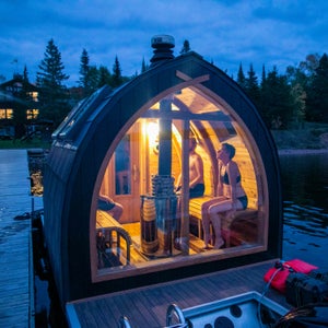 Light from a small floating sauna shines out against wooded lake at dusk.