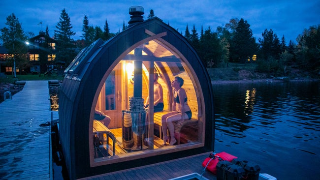 Light from a small floating sauna shines out against wooded lake at dusk.