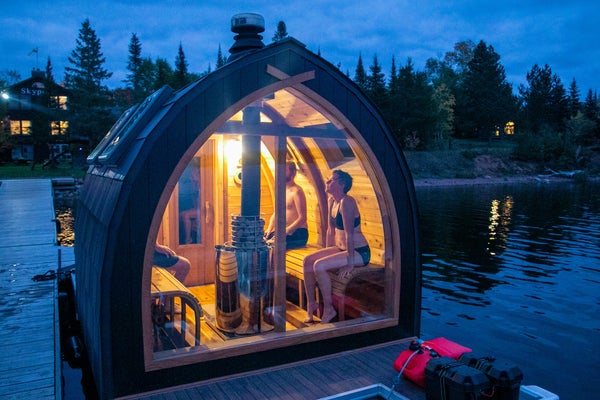 Light from a small floating sauna shines out against wooded lake at dusk.