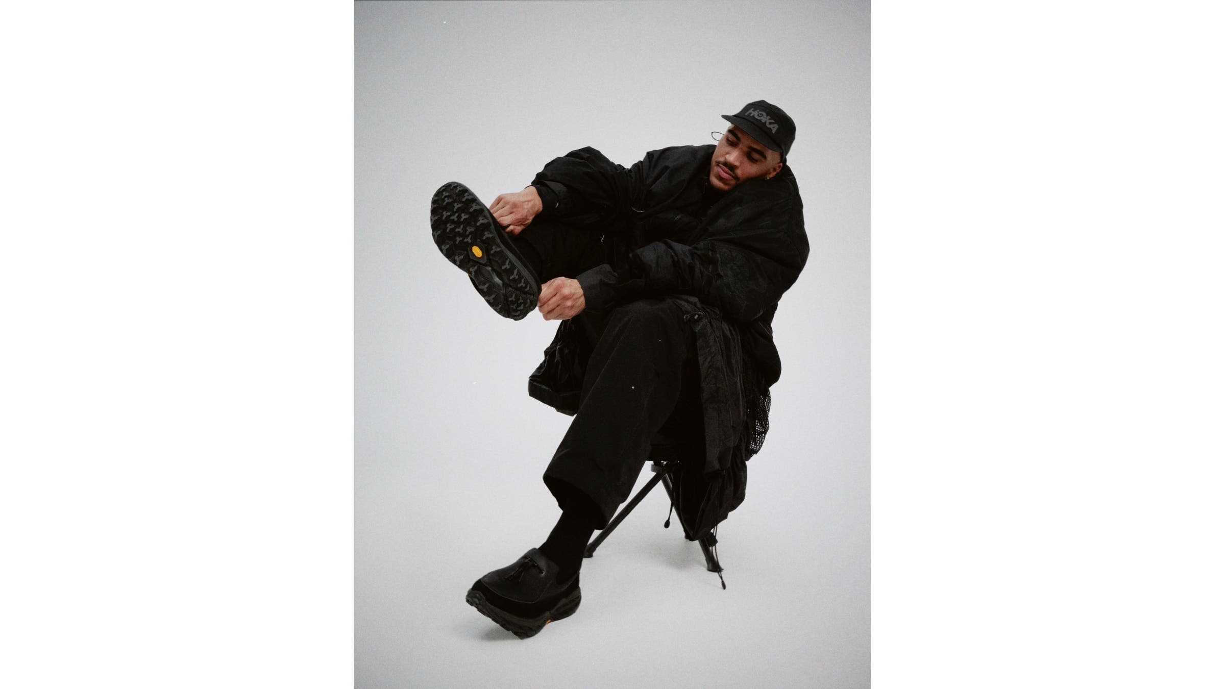 A man sitting in a chair, wearing all black and pulling on black snoafers.