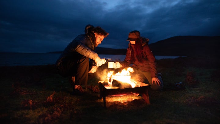 Two people enjoy whiskey over a campfire at night.