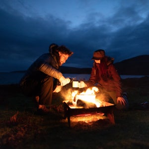 Two people enjoy whiskey over a campfire at night.