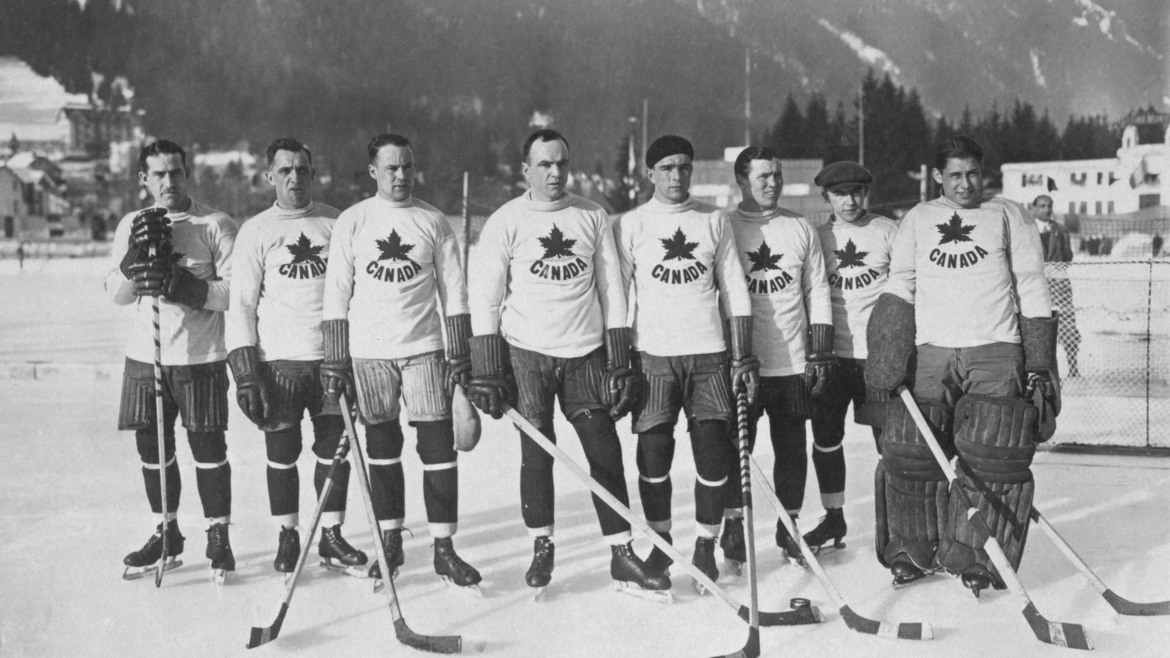 The Toronto Granites hockey team poses on an outdoor rink in their Winter Olympics gear from 1924, featuring wool sweaters emblazoned with "CANADA" and a maple leaf, paired with soft headwear like toques and berets instead of helmets.