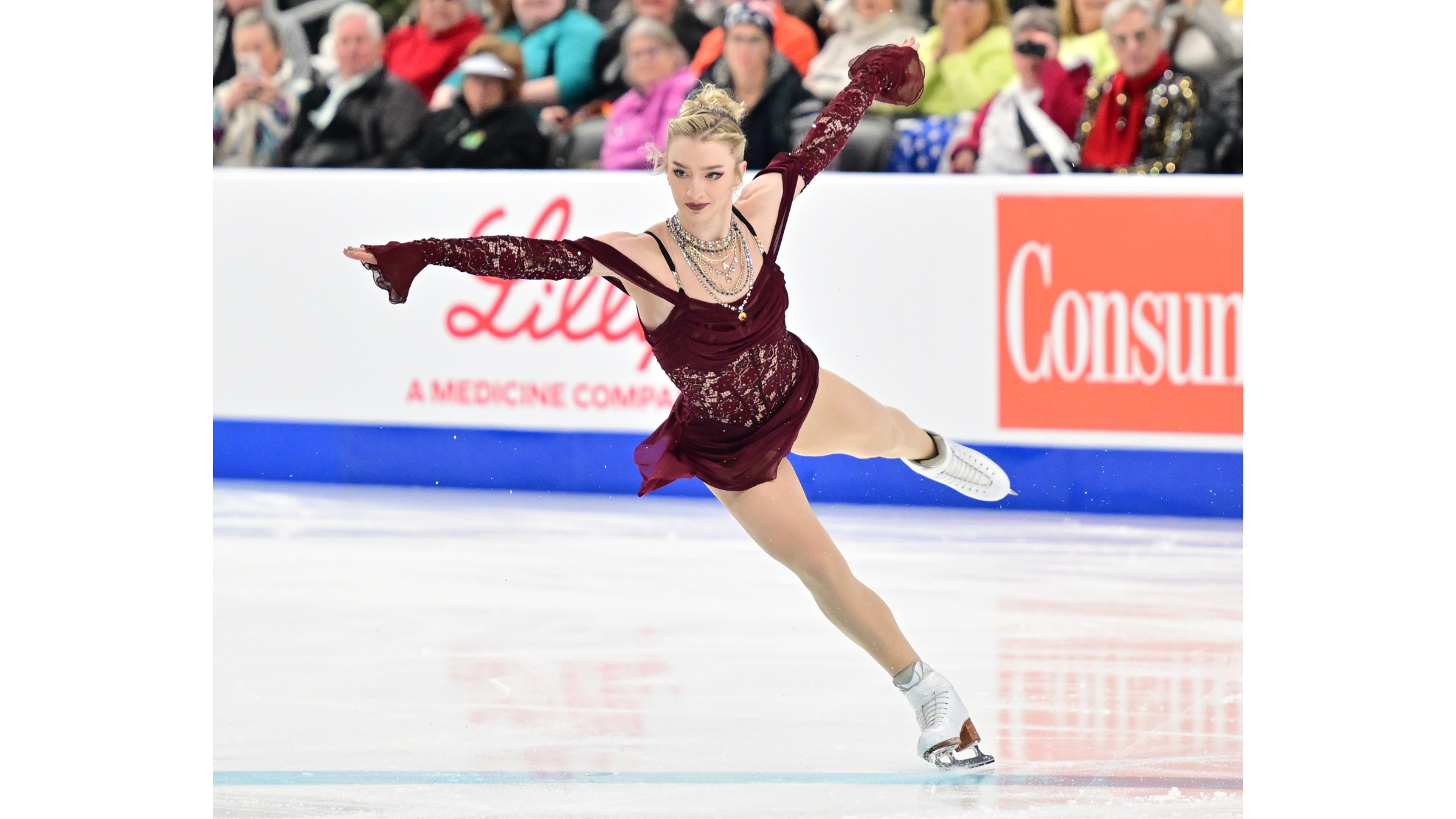 Figure skater Amber Glenn glides across the ice in a modern example of winter olympics gear, wearing a detailed burgundy lace corset dress with matching long sleeves and a multi-strand gold sequined necklace.