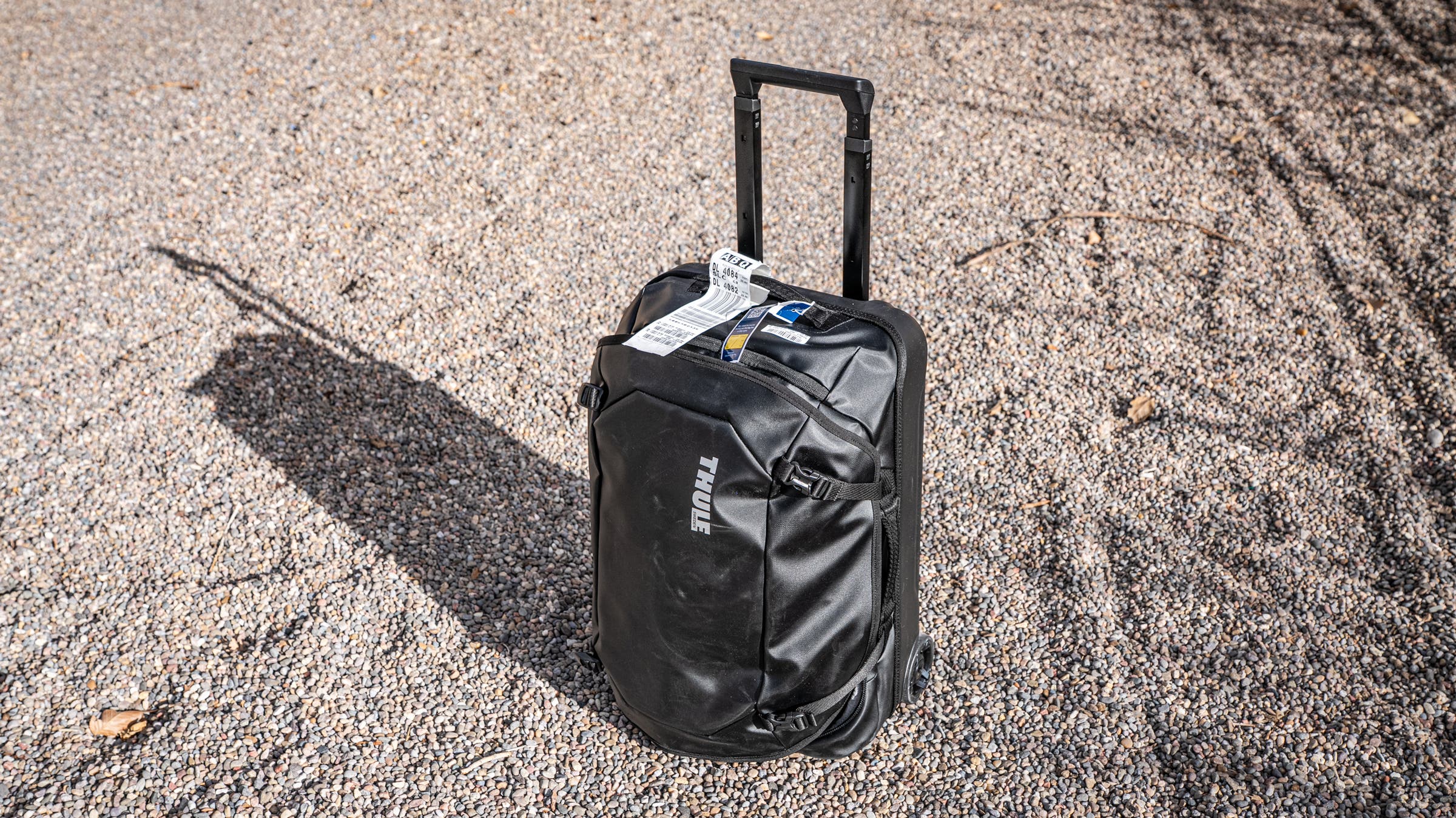 A black Thule Chasm rolling duffel stands upright on a gravel surface with its telescoping handle extended, showing a durable, weather-resistant travel bag designed for rugged trips and gear-heavy travel.