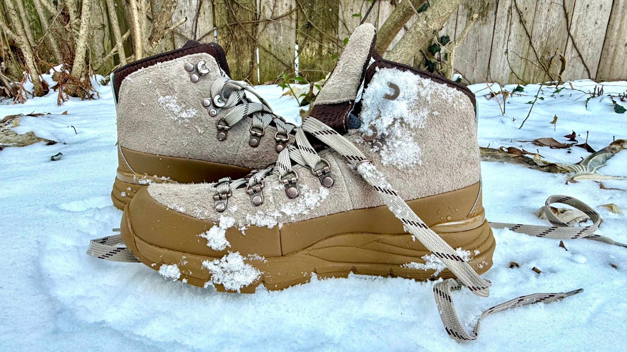 A pair of winter hiking boots dusted with fresh snow, shown side by side outdoors, featuring the suede-and-leather Danner 600 ID Gore-Tex boots with rugged soles and metal lace hooks, photographed in a snowy backyard setting.