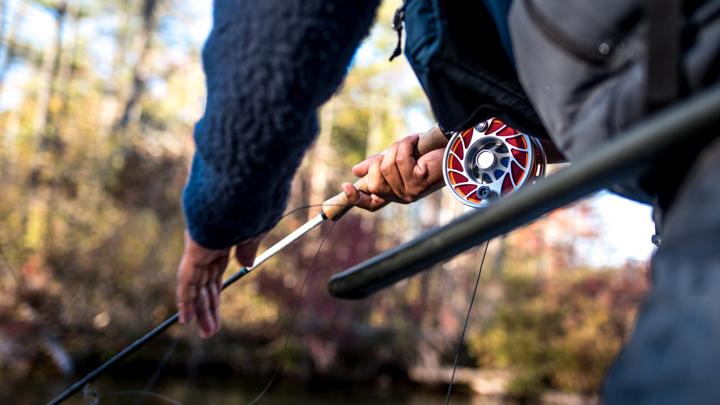 Angler holding a fishing rod and reel while fly fishing along a wooded stream, with a close-up of the reel and cork handle in natural light, photographed by Brent Doscher.