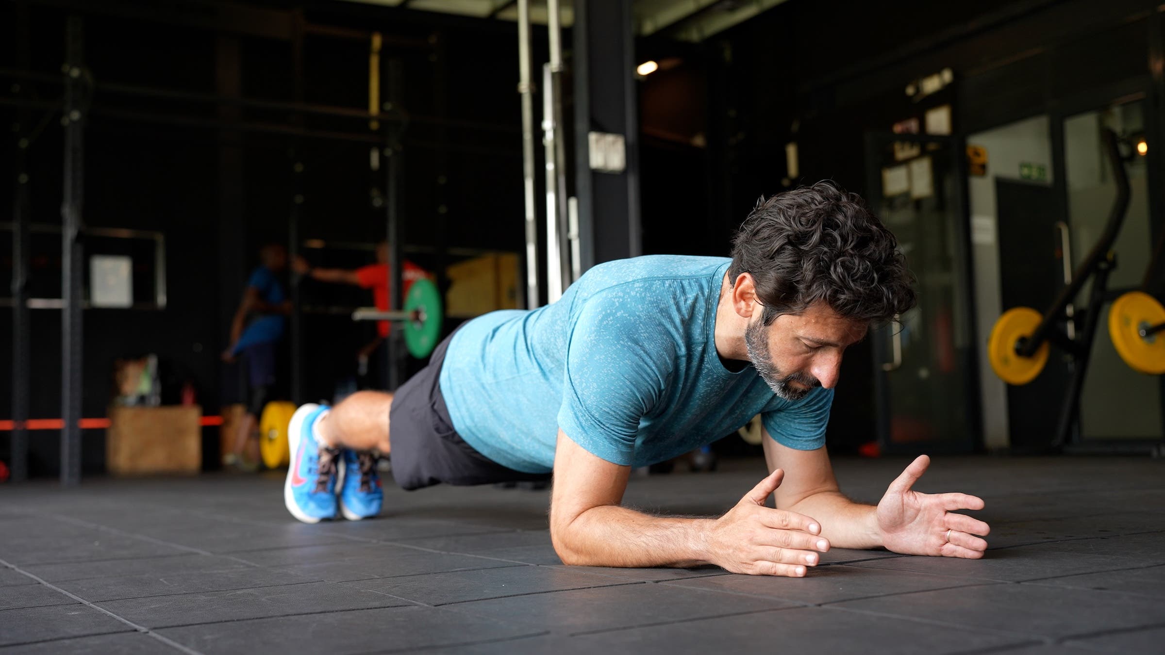 a man with dark hair wearing a blue shirt and black shorts demonstrates how to do a forearm plank for longevity