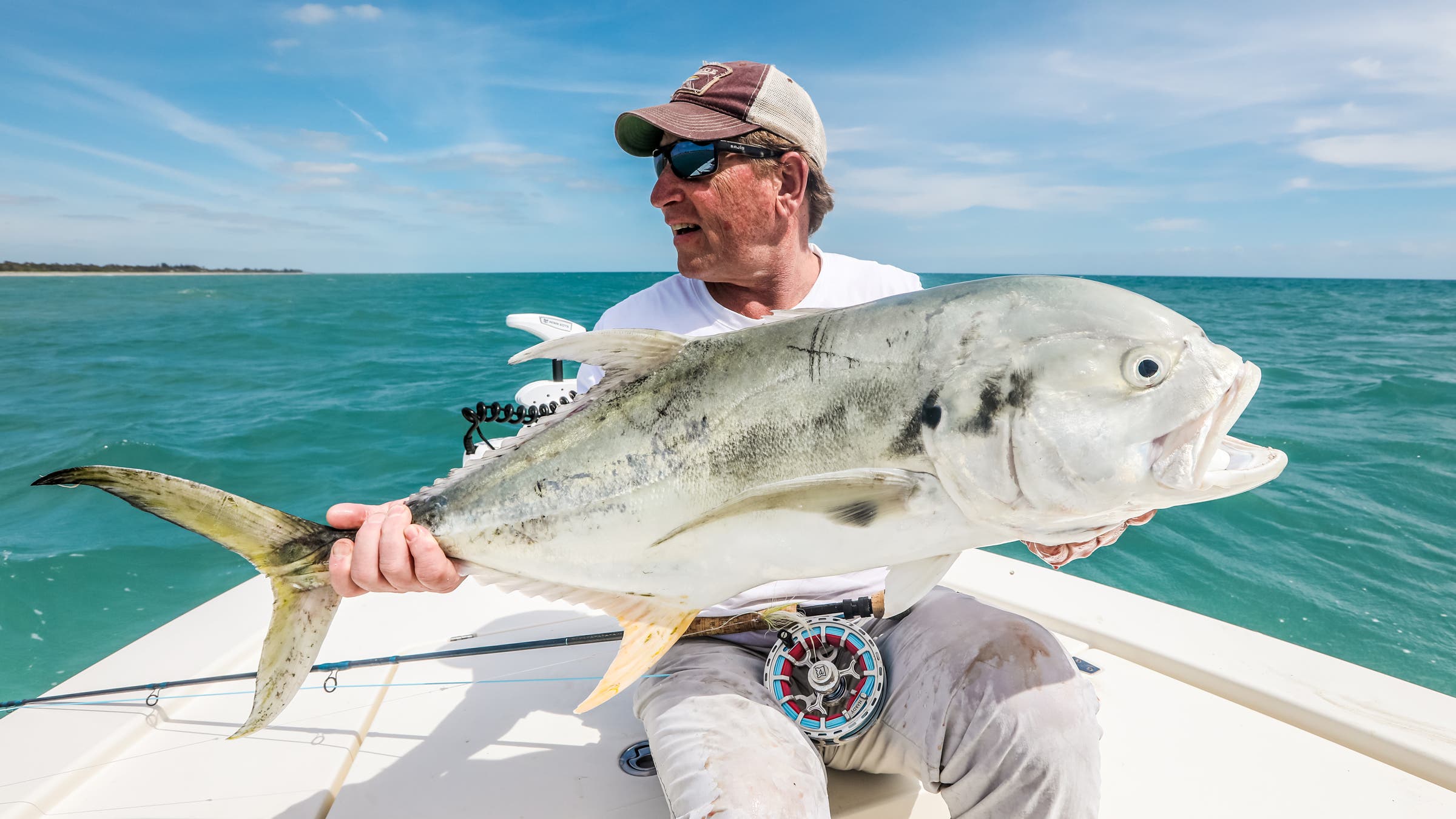 A triumphant angler sits on a boat holding a large Jack Crevalle caught while saltwater fly fishing. The image prominently displays a high-performance fly reel in the foreground with the bright turquoise sea in the background.