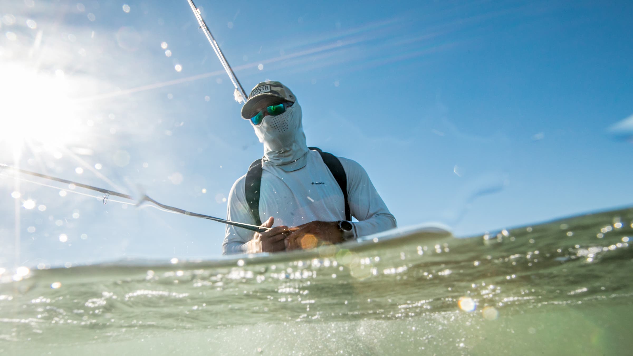 An angler wading deep in the ocean wears a sun mask and polarized sunglasses for protection while saltwater fly fishing in bright, harsh conditions.