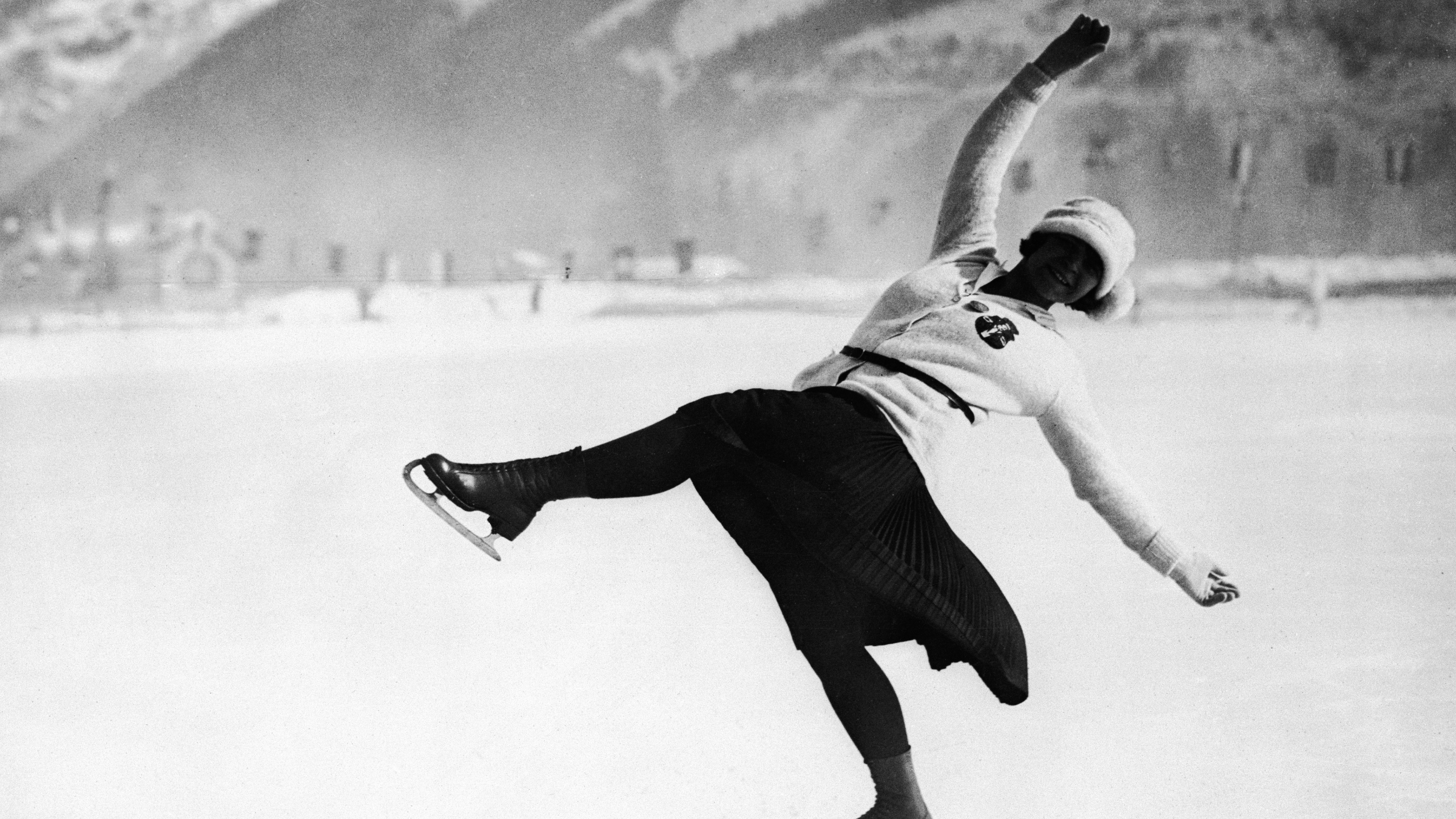 A black and white historical photograph of figure skater Herma Szabo performing a dynamic one-legged glide on an outdoor ice rink. She is wearing a 1920s-style belted cardigan, a calf-length pleated skirt, and a cloche hat against a snowy mountain backdrop.