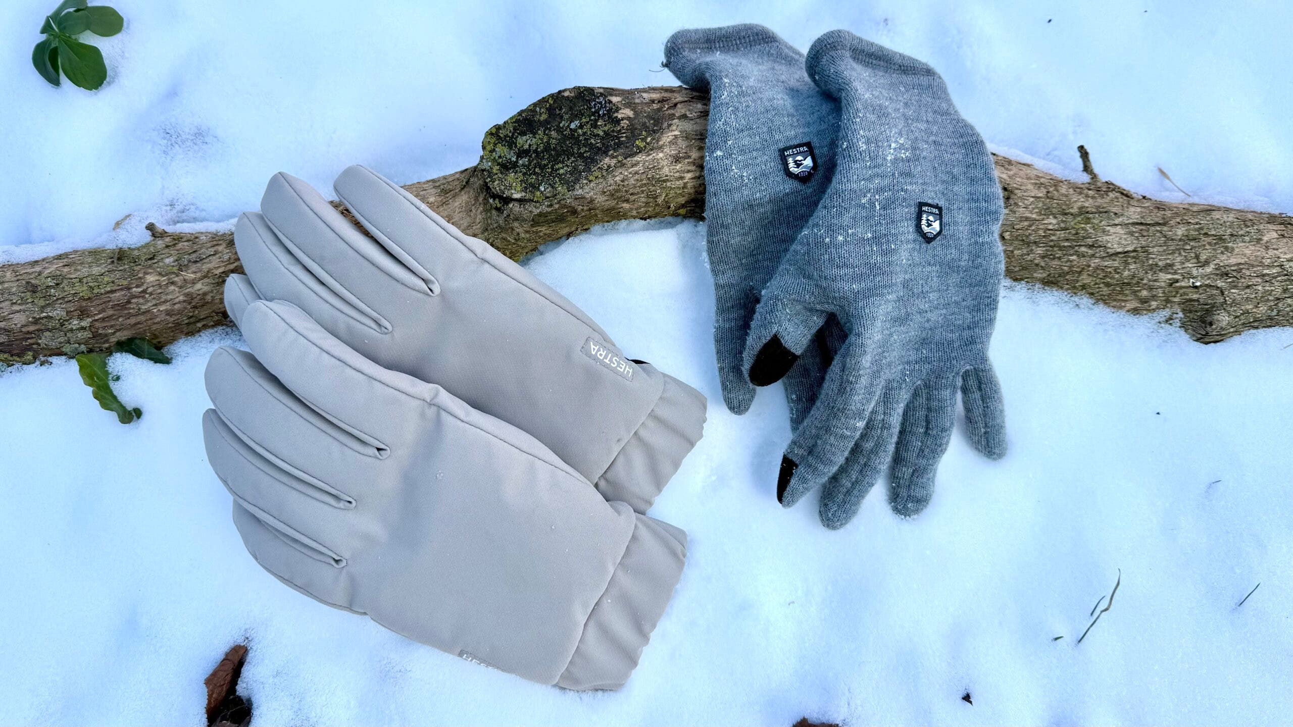 A flat lay of winter gloves arranged on fresh snow, showing the insulated Hestra Axis Gloves alongside the Hestra Gravita Merino Liner gloves draped over a fallen branch, highlighting a layered handwear system for cold-weather conditions.