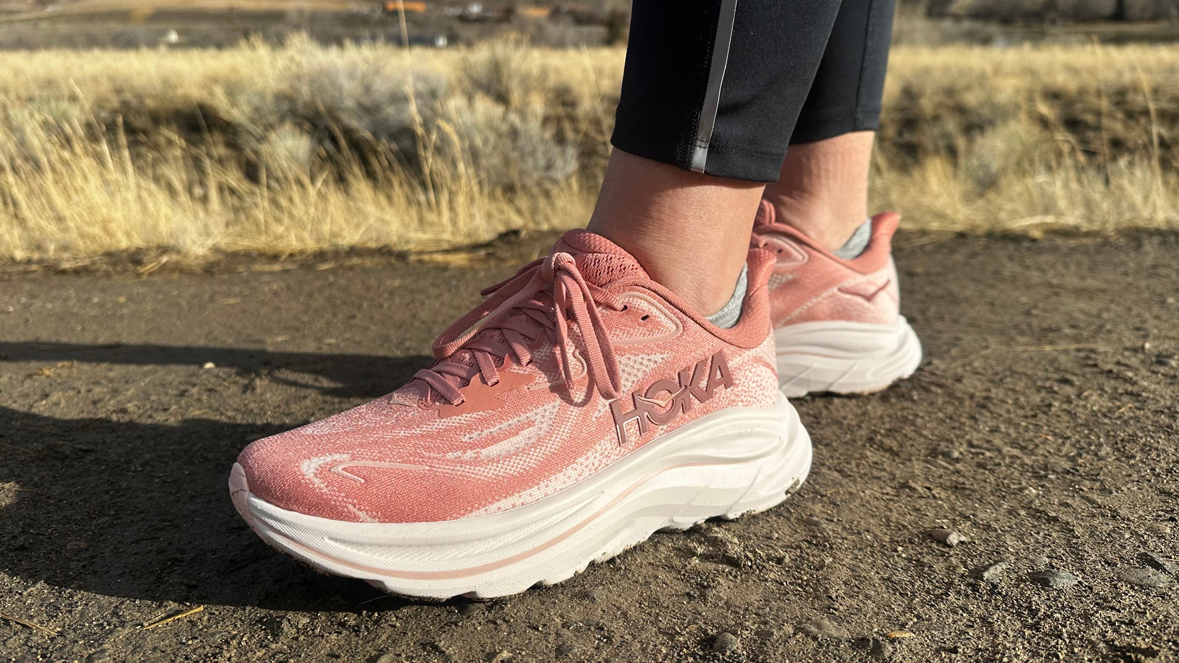 Close-up of Hoka Clifton 10 running shoes on a dirt path, highlighting the plush cushioned midsole, breathable mesh upper, and everyday trainer design for road running.