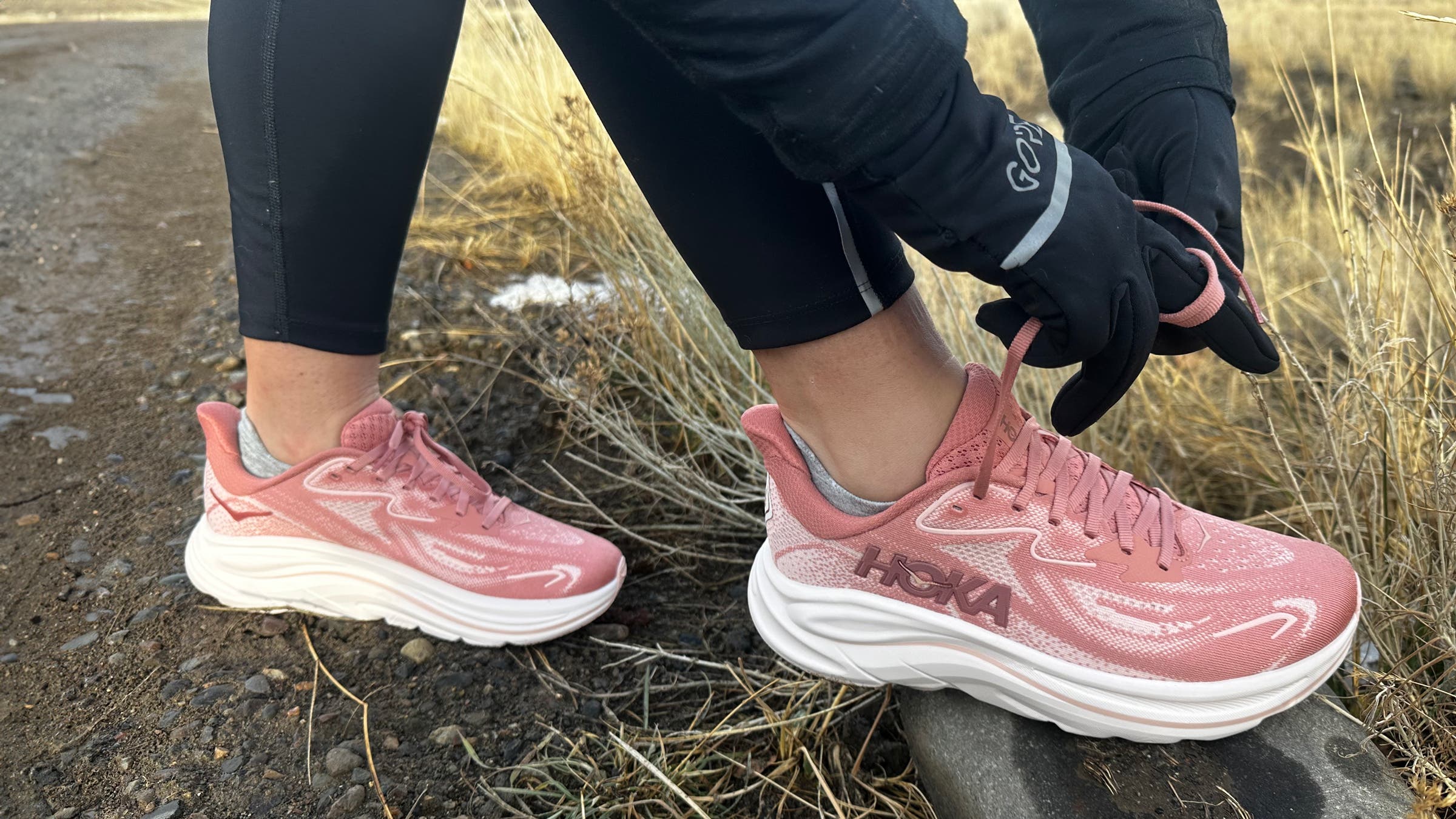 runner ties the laces on pink Hoka Clifton 10 running shoes beside a dirt path, highlighting the cushioned midsole and everyday training fit for outdoor miles.