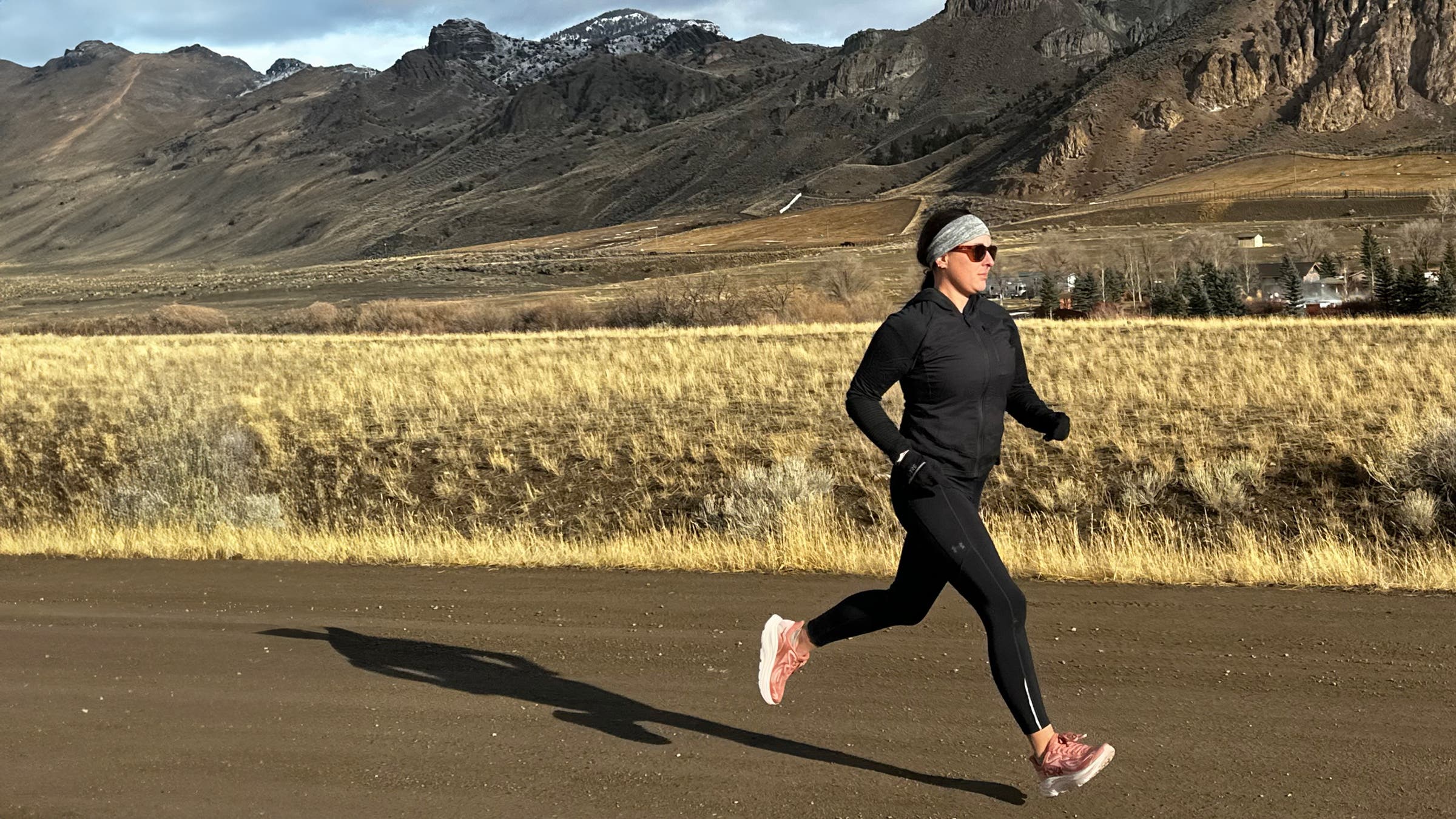 Author and running shoe tester, Maggie Slepian, moves along a dirt road through an open valley with rocky mountains in the background, wearing black layers and cushioned running shoes during an outdoor training run.