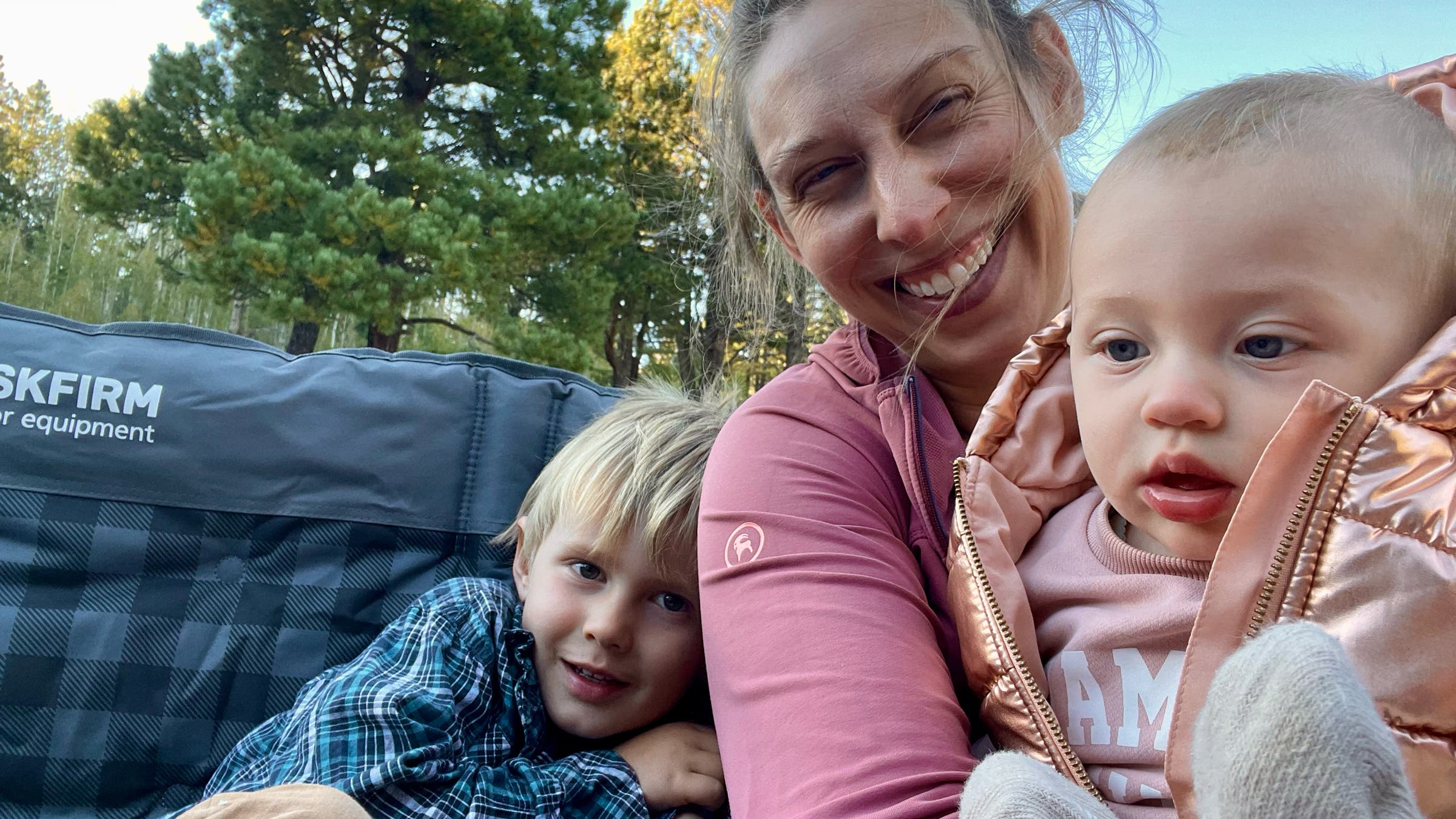 Abigail Wise relaxes with her family in a forest campsite, sharing a cushioned loveseat camping chair that offers enough space for an adult and two young children to sit comfortably outdoors.