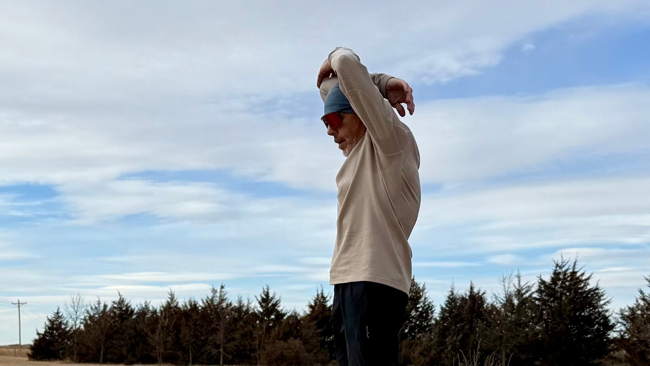 A runner stretches with arms overhead in an open field while wearing the Janji Waffleloft Longsleeve, highlighting the breathable, textured fabric during a cool-weather workout.