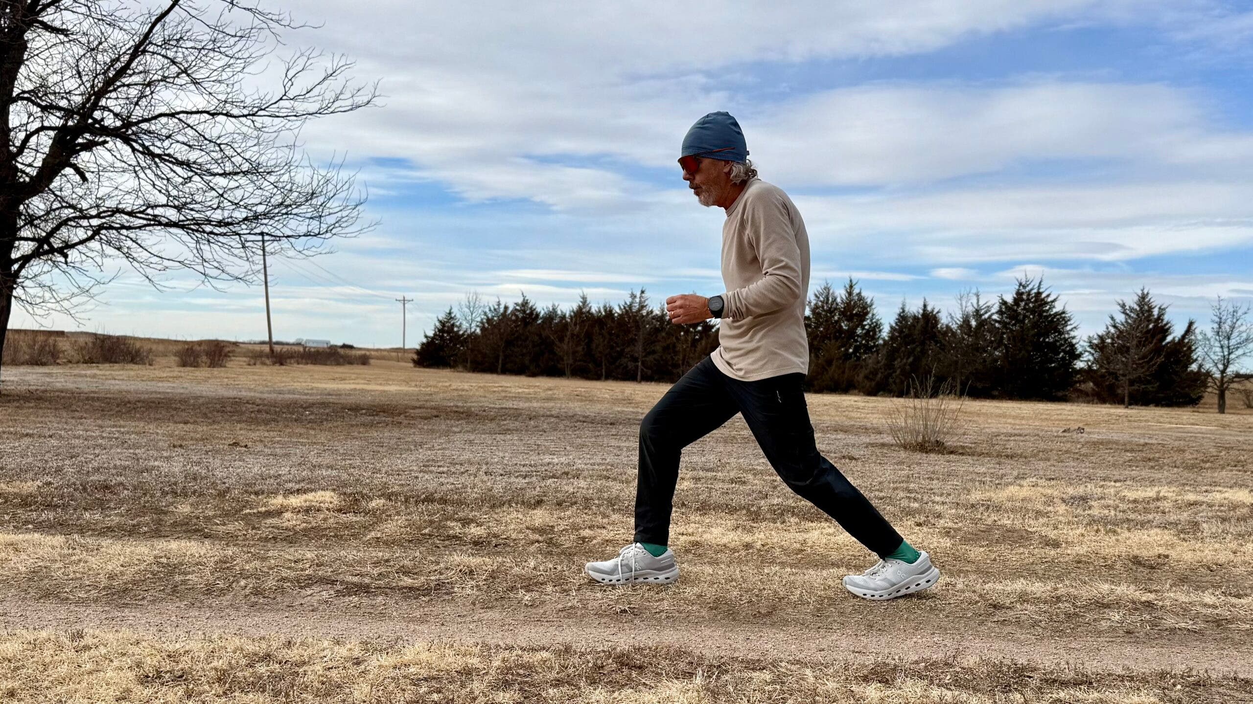 A runner moves along a dirt path in an open field wearing the Janji Waffleloft Longsleeve, showing the lightweight, textured top in motion during a cool-weather run.