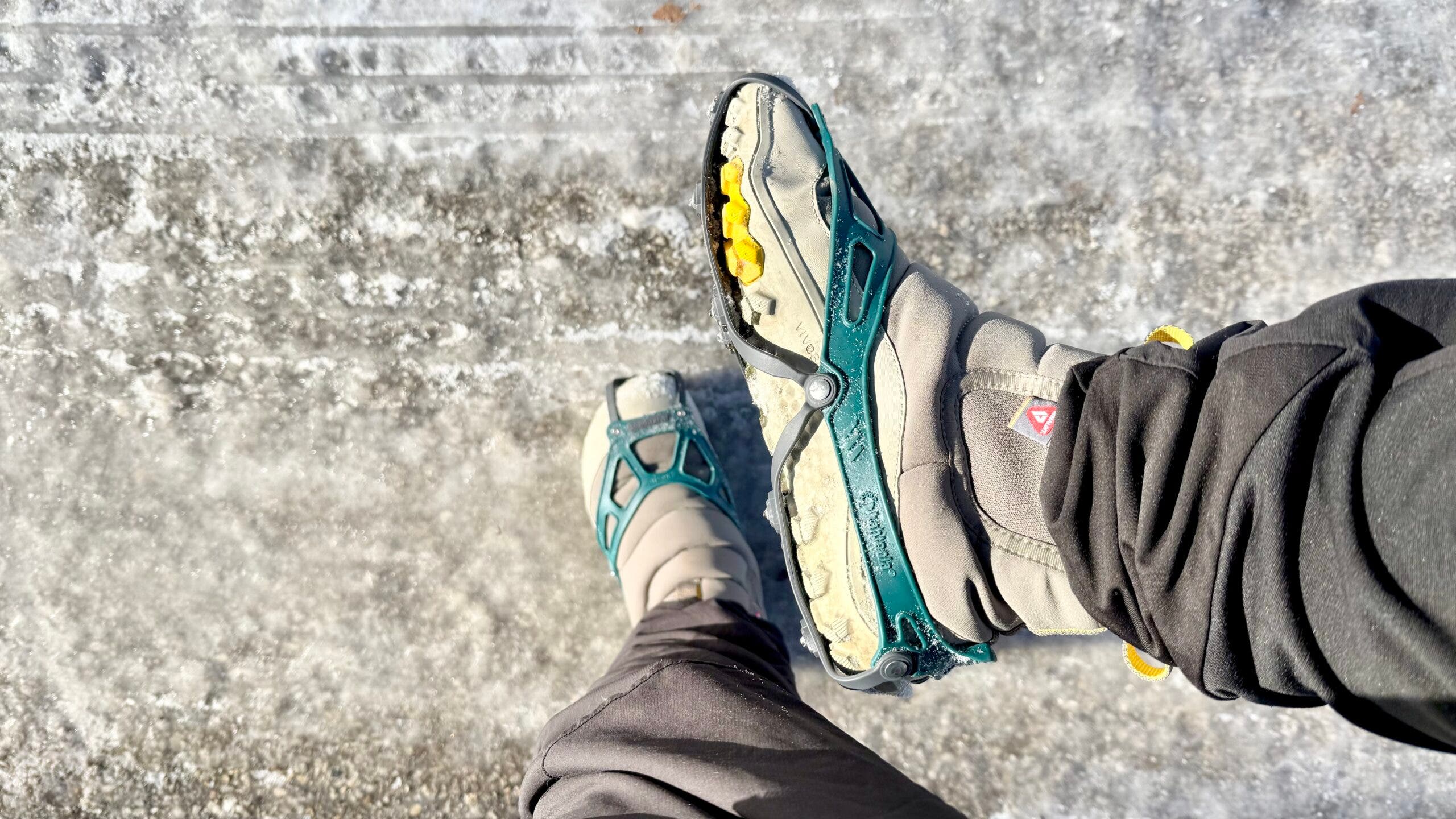 A close-up of boots stepping across icy pavement while wearing winter crampons, showing Kahtoola NANOspikes fitted over Vivobarefoot Explorer JJF boots, with metal studs gripping slick, frozen ground during urban winter walking conditions.