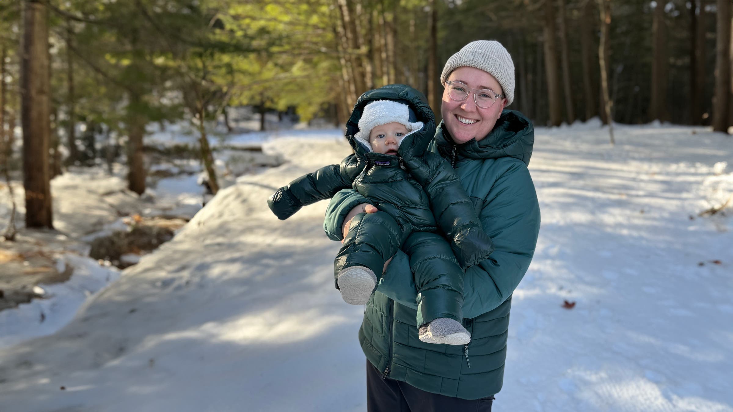 Contributor Erica Zazo holds a bundled baby on a snowy forest trail, with the child wearing the Patagonia Infant Hi-Loft Down Sweater Bunting, a puffy insulated baby jacket designed to keep infants warm during winter outings.