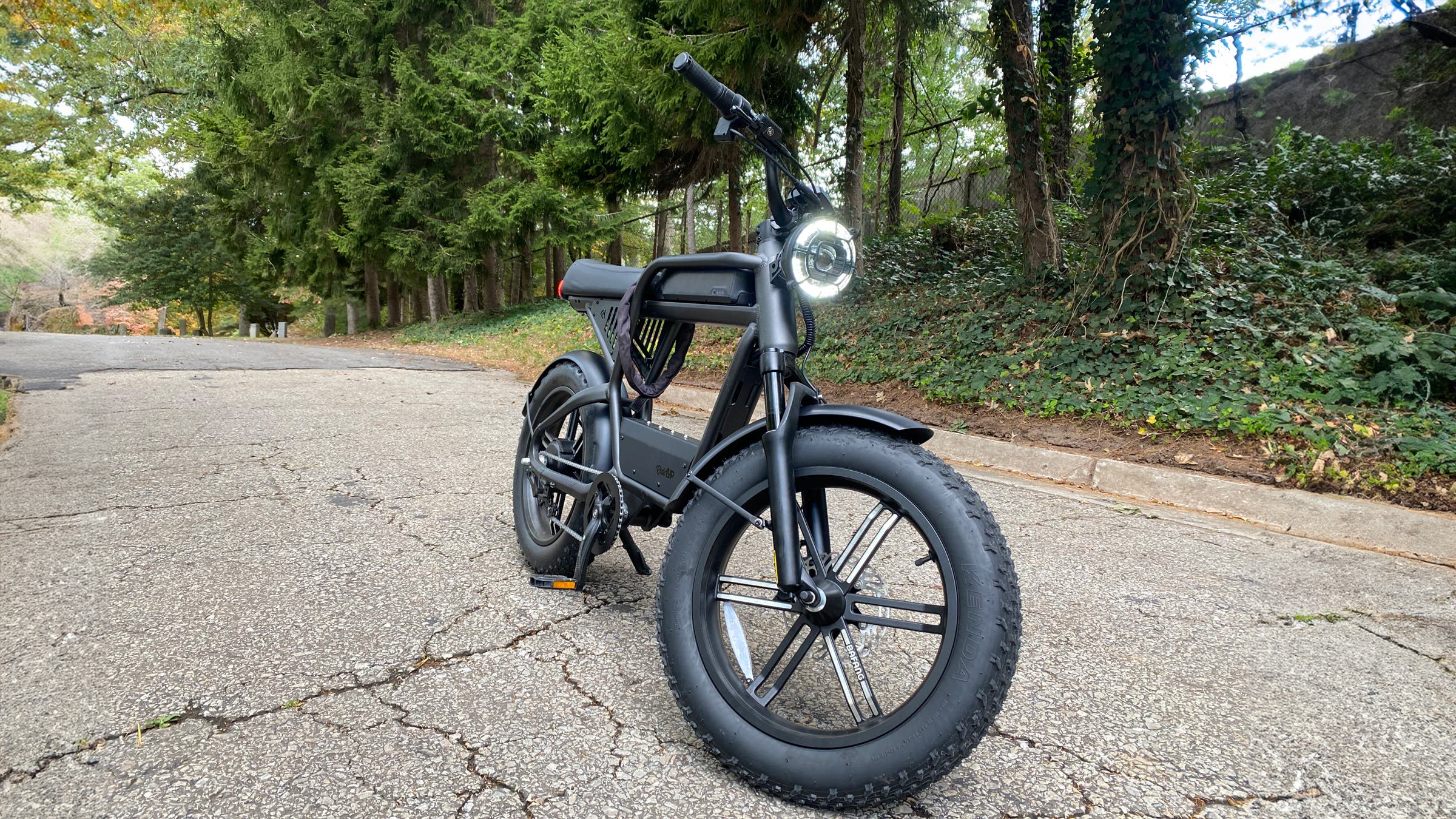 A black Ride1Up Revv1HT electric bike with fat tires is parked on a quiet, tree-lined road, showcasing its moto-inspired frame, integrated headlight, and rugged design built for urban streets and mixed terrain.