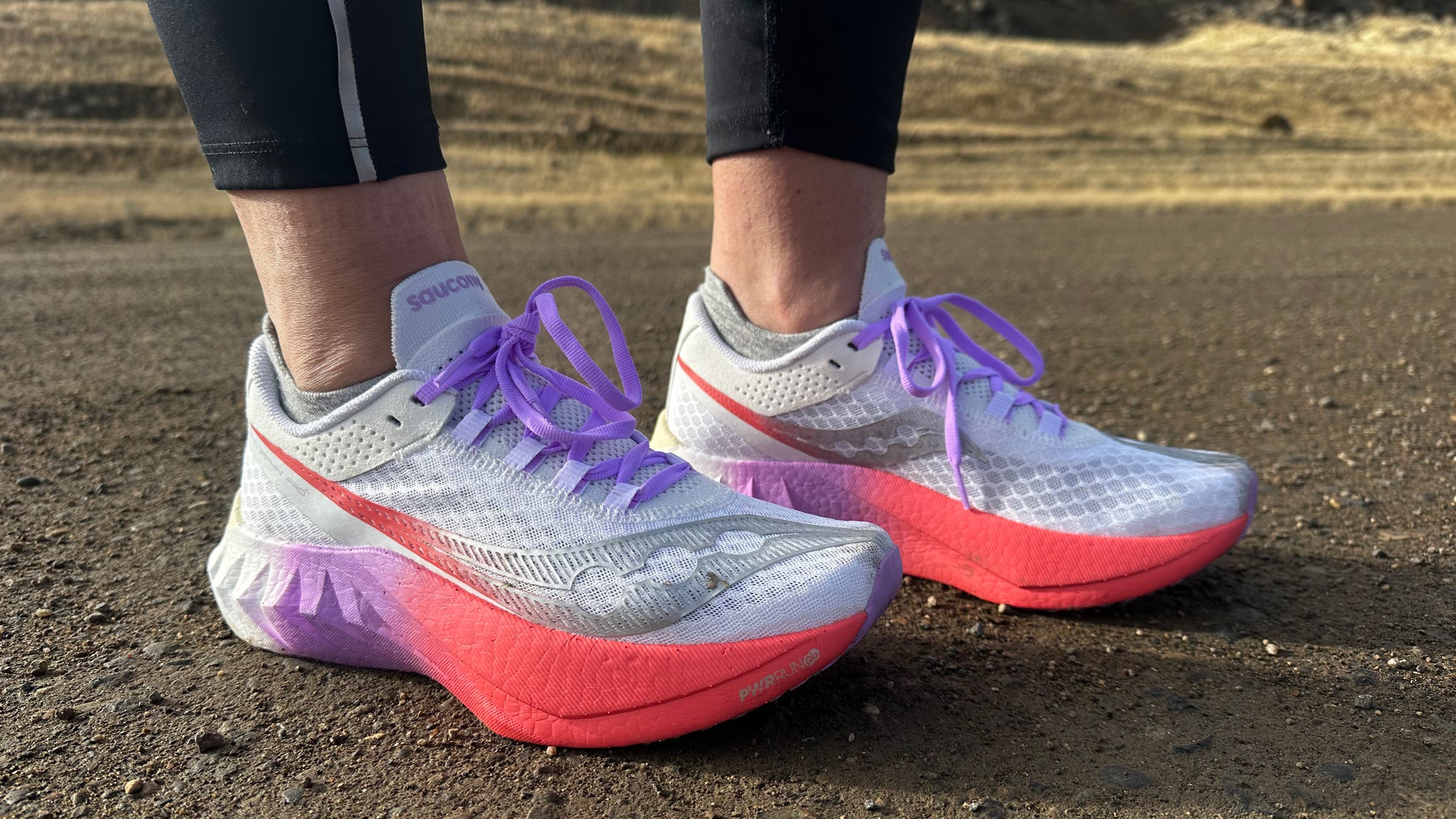 Close-up of Saucony Endorphin Pro 4 ($240) running shoes on a gravel road, showing the lightweight mesh upper, purple laces, and high-stack cushioned midsole designed for speed and performance.