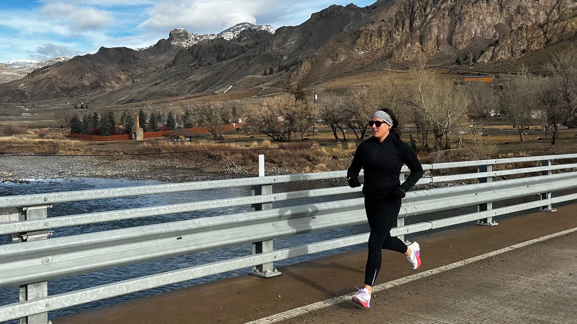 A runner jogs across a bridge beside a river with rocky mountains in the background, wearing black layers and bright running shoes during an outdoor training run.