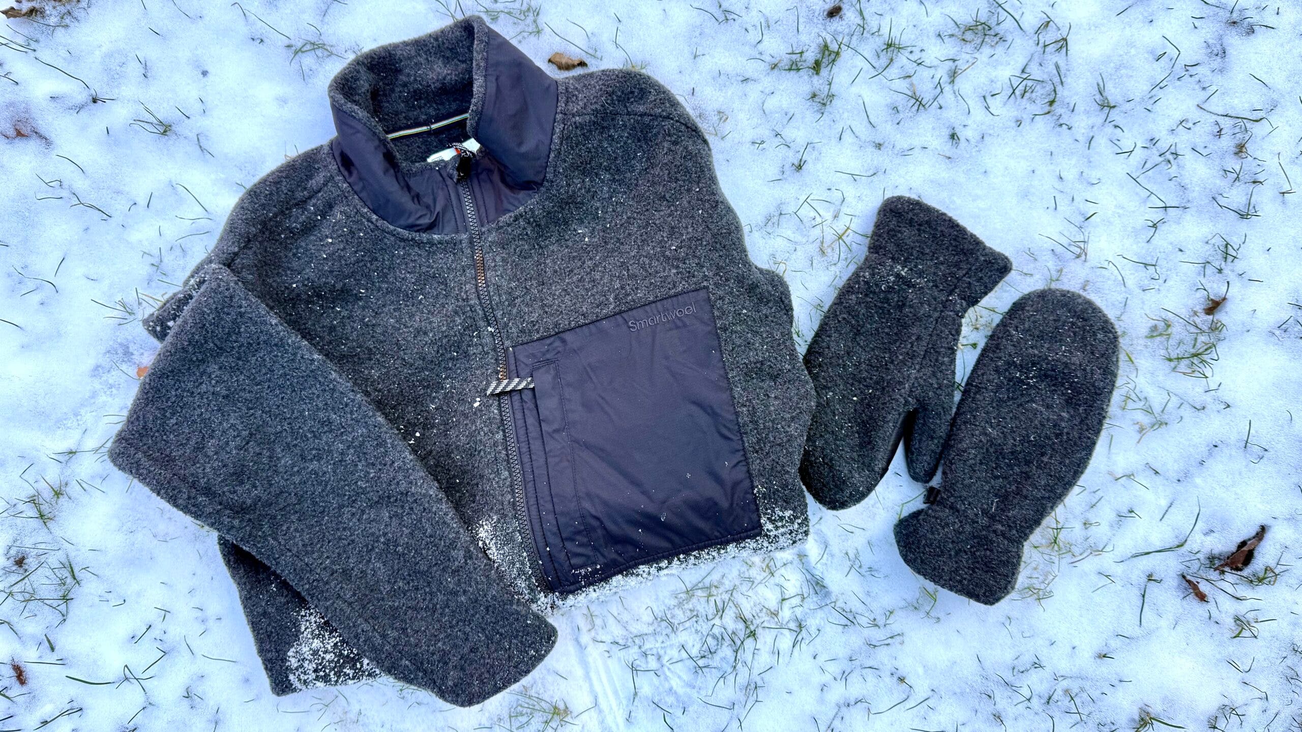 A winter fleece jacket and gloves resting on snow, featuring the Smartwool Second Cut fleece and gloves, lightly dusted with snow to highlight a cozy, cold-weather layering setup.