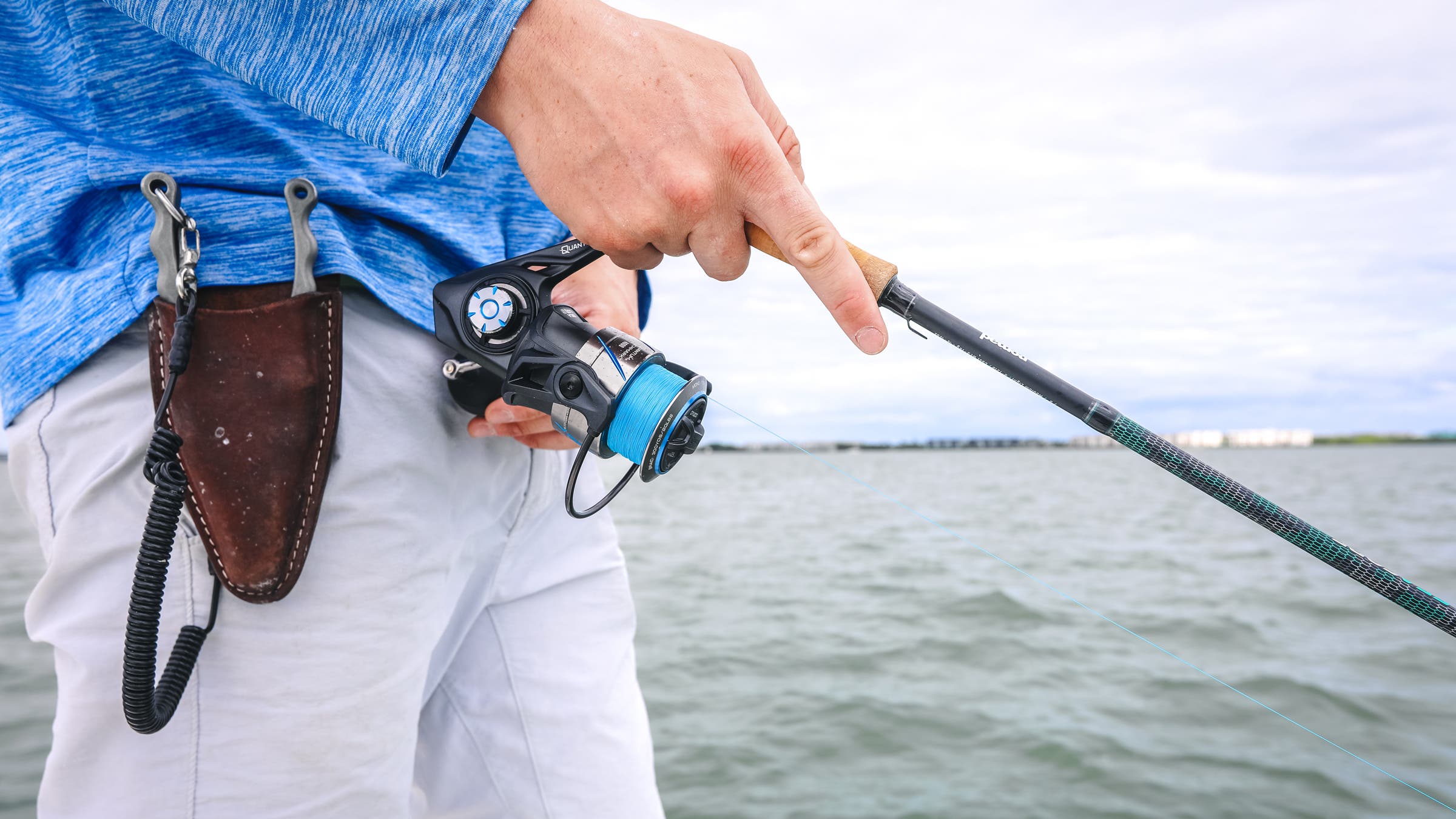 A close-up of an angler holding a fishing rod and reel while out saltwater fly fishing, featuring high-end pliers in a leather holster at his waist for quick access. The scene is set against a vast body of water under a cloudy sky.