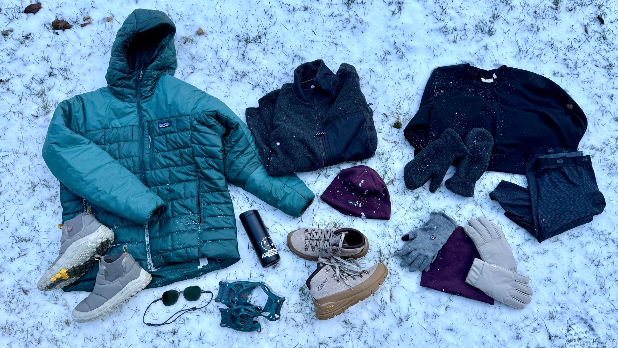 A flat lay of cold-weather essentials arranged on fresh snow, including an insulated teal puffer jacket, fleece layers, winter boots, mittens, gloves, hats, microspikes, sunglasses, and a water bottle—gear curated for urban winter conditions in a story by author Erica Zazo.