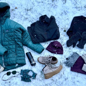 A flat lay of cold-weather essentials arranged on fresh snow, including an insulated teal puffer jacket, fleece layers, winter boots, mittens, gloves, hats, microspikes, sunglasses, and a water bottle—gear curated for urban winter conditions in a story by author Erica Zazo.