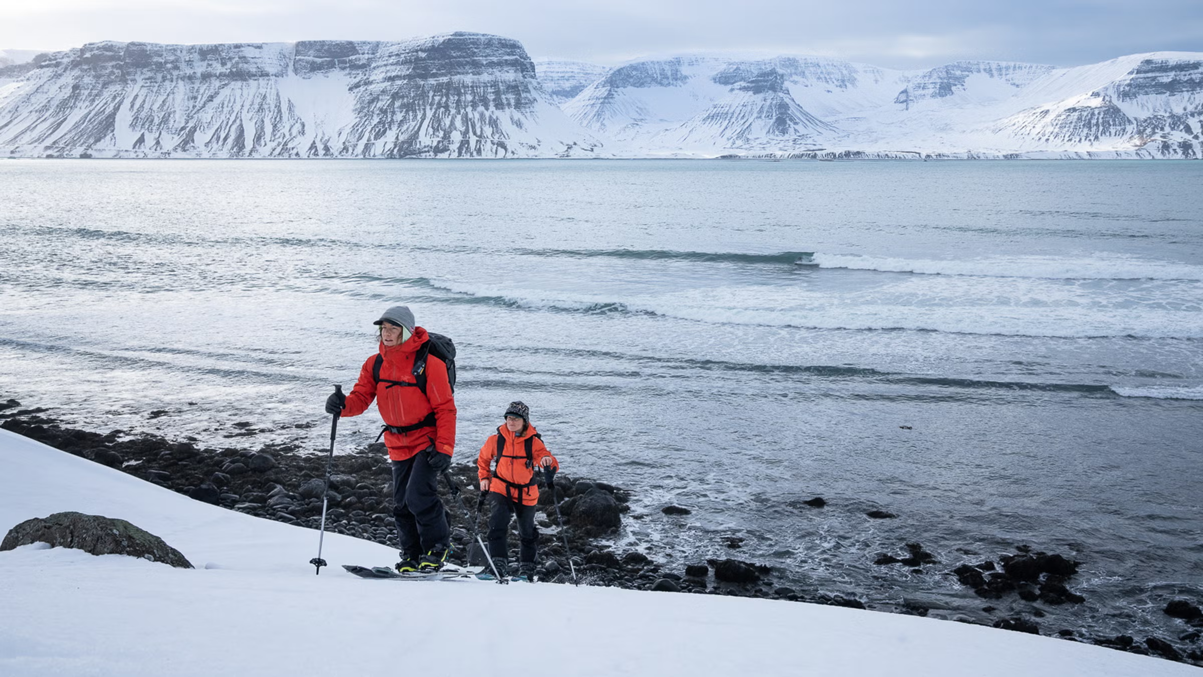 Two hikers wearing red and orange Arc’teryx shells and backpacks trek through a snowy coastal mountain range.