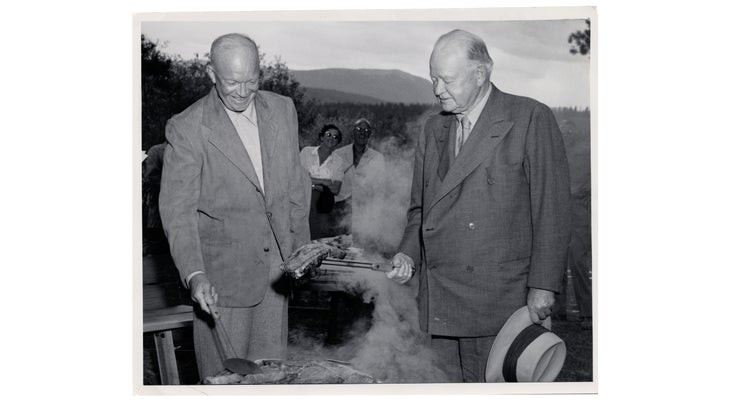Black and white photo of President Eisenhower cooking a dirty steak while others look on.