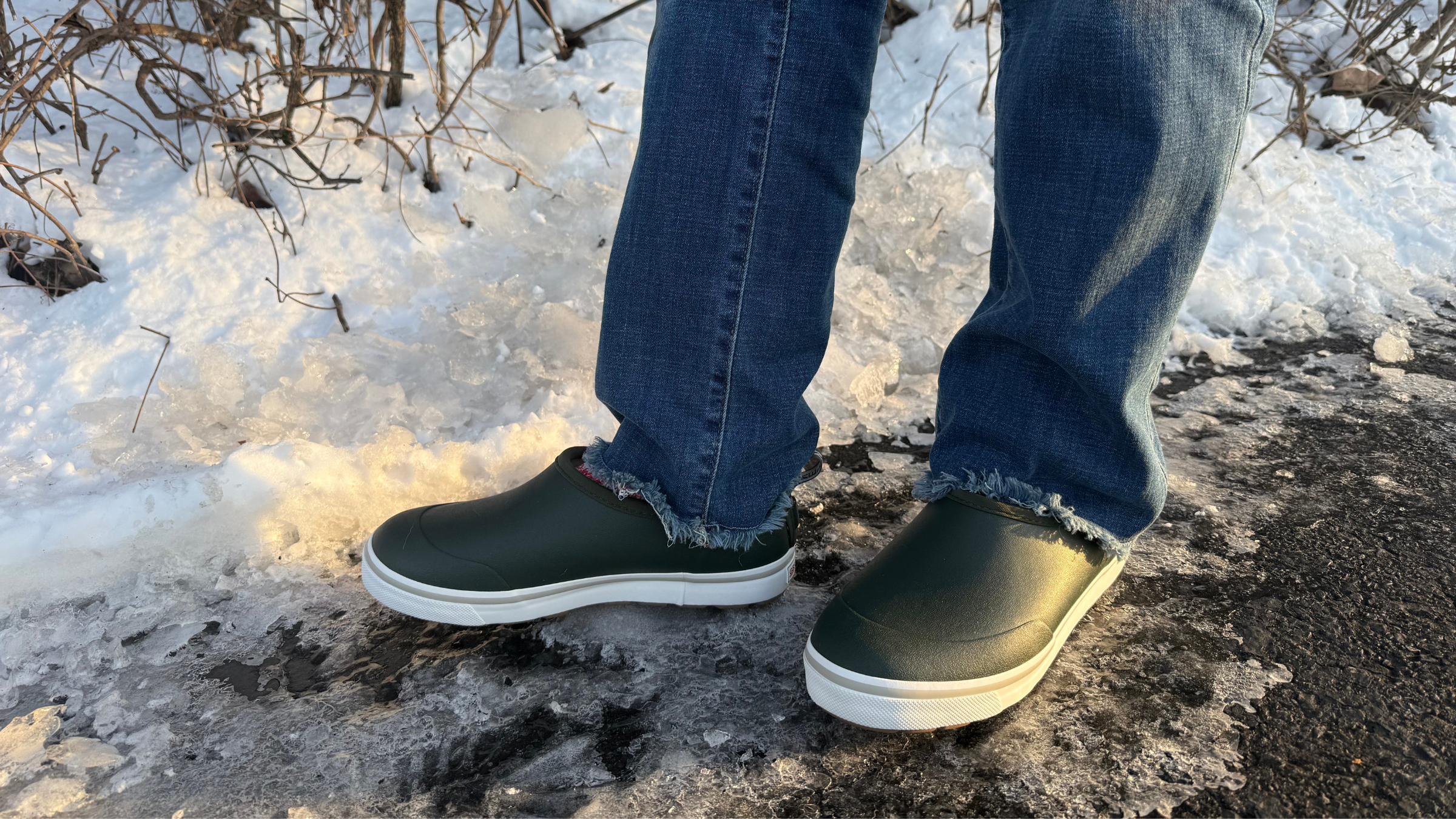 A person wearing a pair of dark green Grundens Deck Boss Shorty shoes and blue jeans, standing on icy ground to demonstrate why they are the best airport shoe for travelers heading into winter weather.