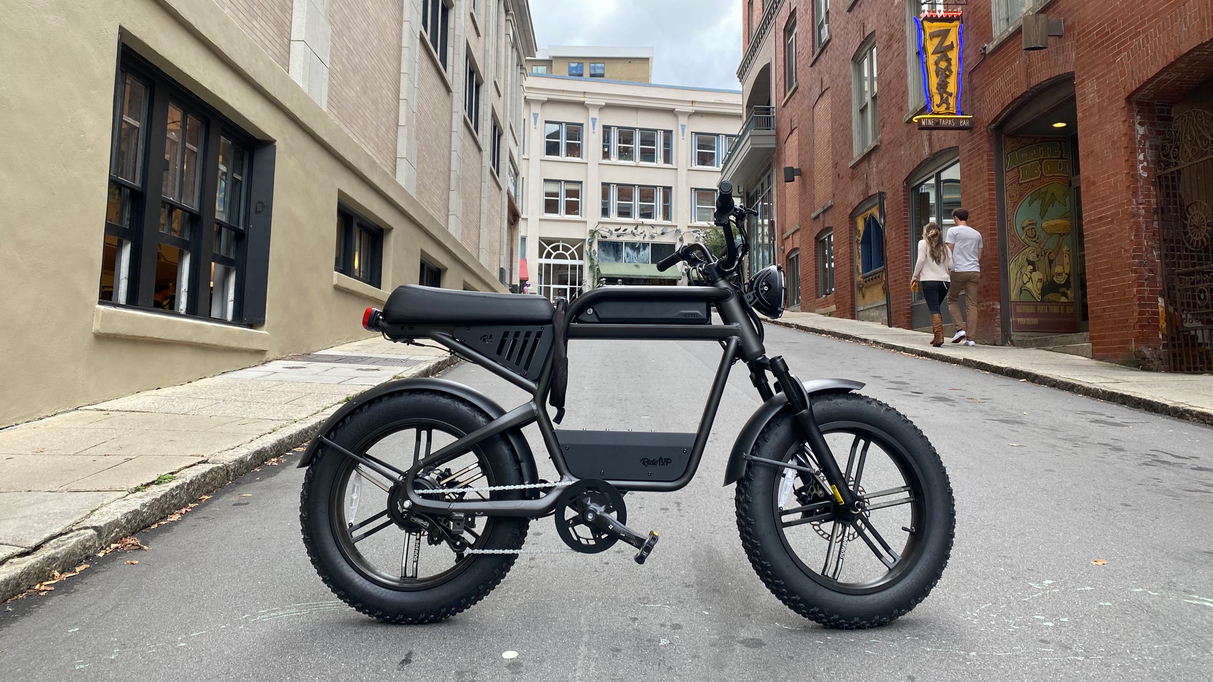 A black Ride1Up Revv1HT electric bike with fat tires is parked in the middle of a narrow downtown street, highlighting its moto-inspired frame, low seat, and rugged urban-ready design.