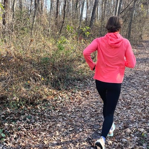 A runner moves along a leaf-covered forest trail wearing a bright pink running midlayer, captured from behind on a wooded path and photographed by author Lisa Jhung.