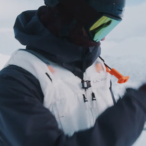 A skier wearing the SBX avalanche survival system integrated into a white vest, showing the orange pull-handle and mouthpiece designed to provide airflow to buried victims.